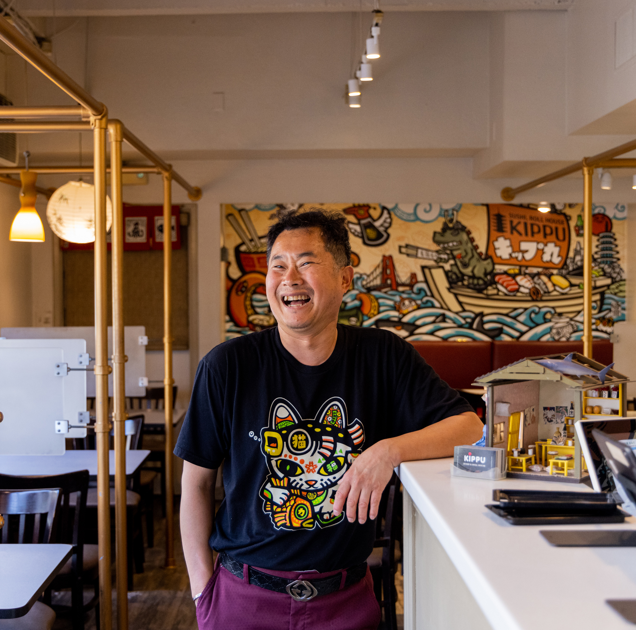 A cheerful man in a black graphic t-shirt stands in a vibrant, colorful restaurant with Japanese-themed decor and murals, smiling broadly.