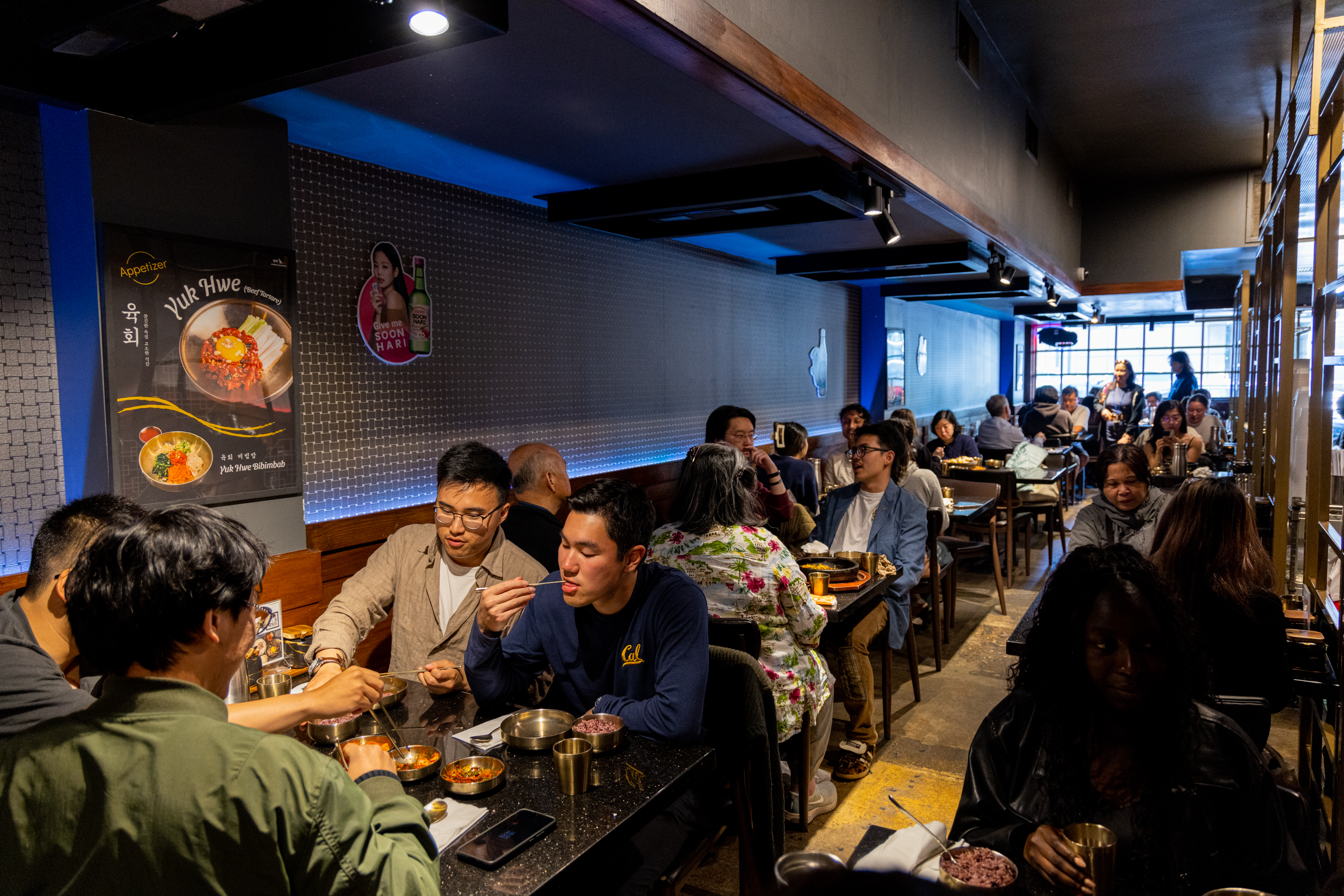 A bustling restaurant with closely packed tables shows diners enjoying their meals. The interior has dim lighting, with posters on the walls and a mix of people conversing.
