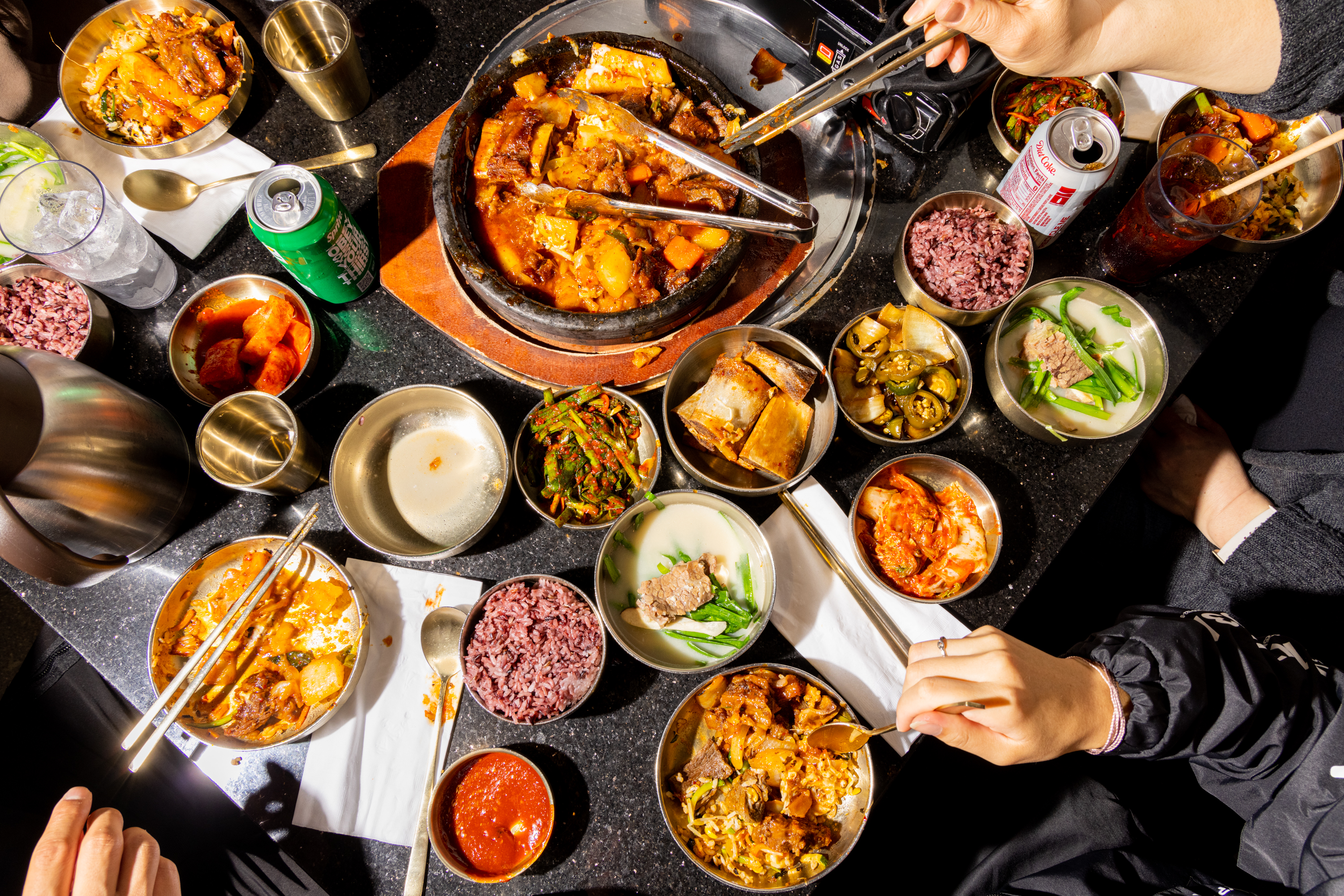 People are sharing a vibrant Korean meal with kimchi, rice, various stews, and side dishes spread out on a table. Drinks include soda cans and water.