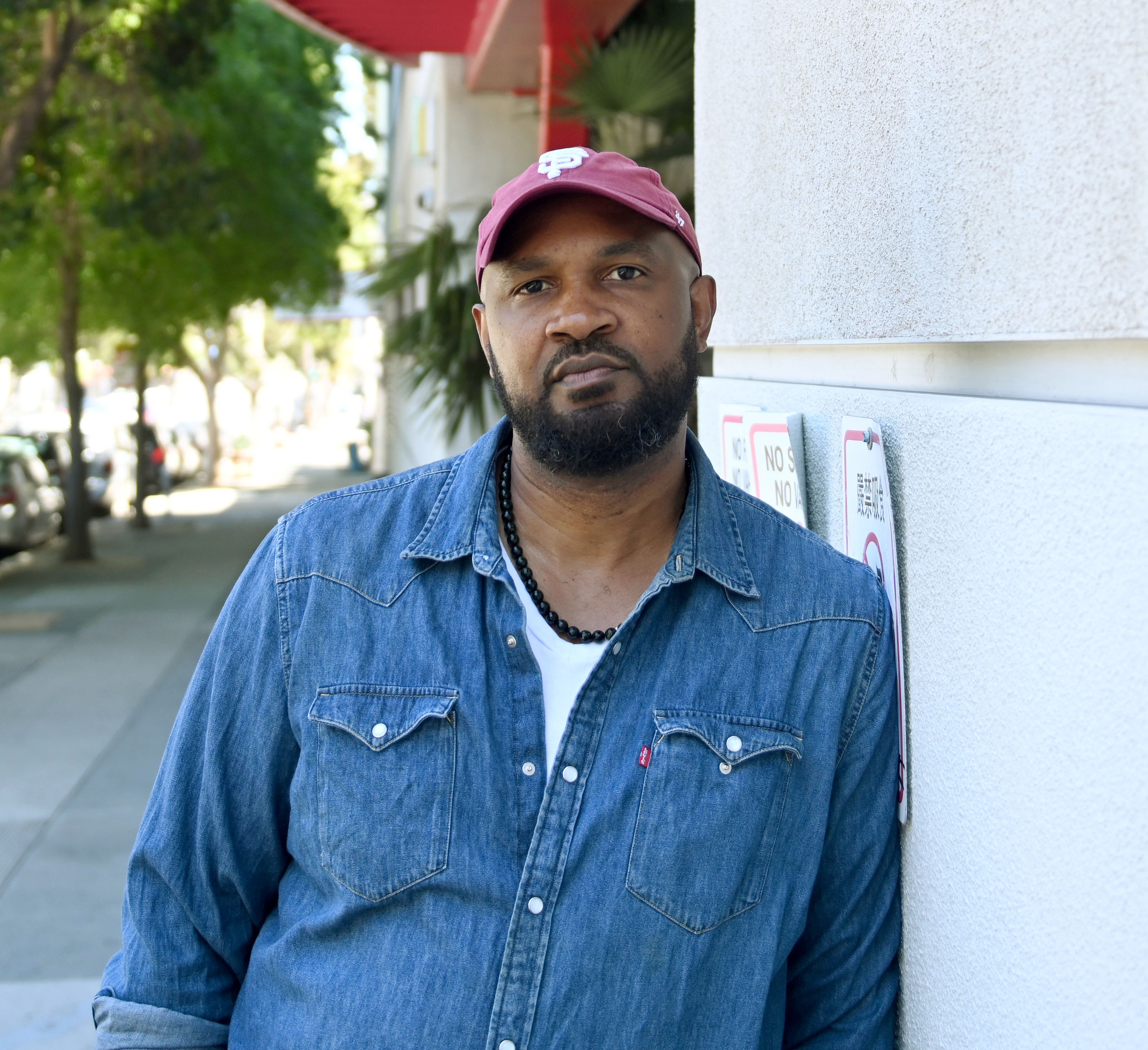 A man with a beard, wearing a red cap, denim shirt, and beaded necklace, leans against a wall on a quiet street lined with trees and parked cars.