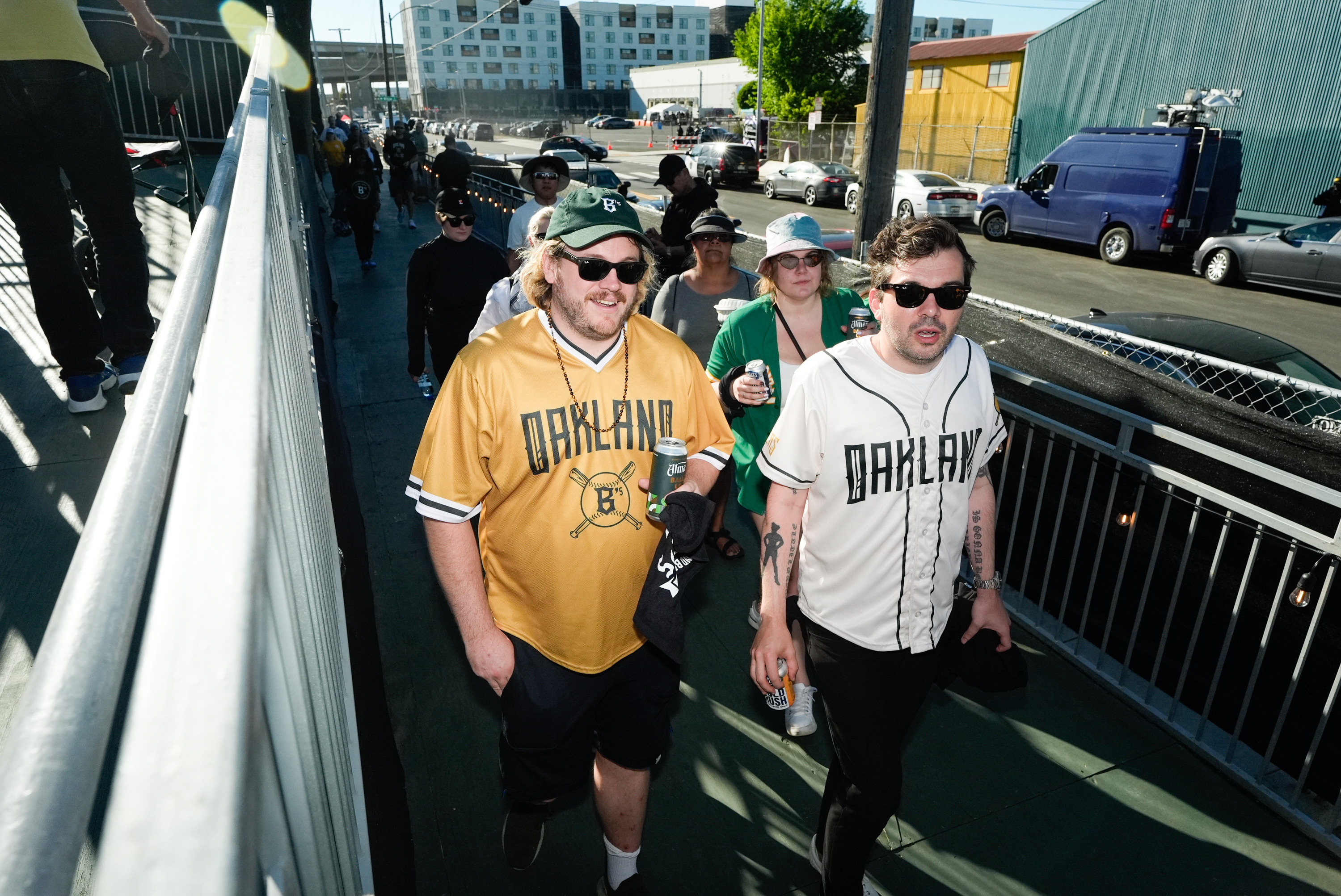 A group of people, most wearing Oakland-themed sports jerseys and sunglasses, are walking up a ramp in an urban outdoor setting on a sunny day.