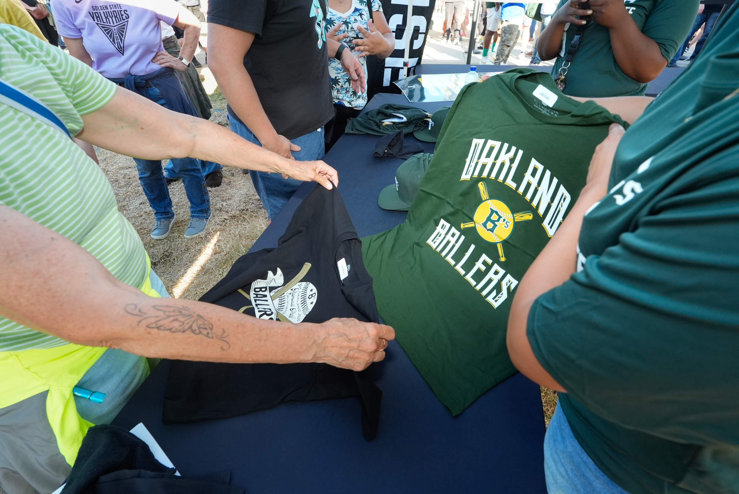 People are gathered around a table, examining green and black shirts with &quot;Oakland Ballers&quot; logos. One person has a tattoo on their arm. Other individuals are wearing printed shirts.