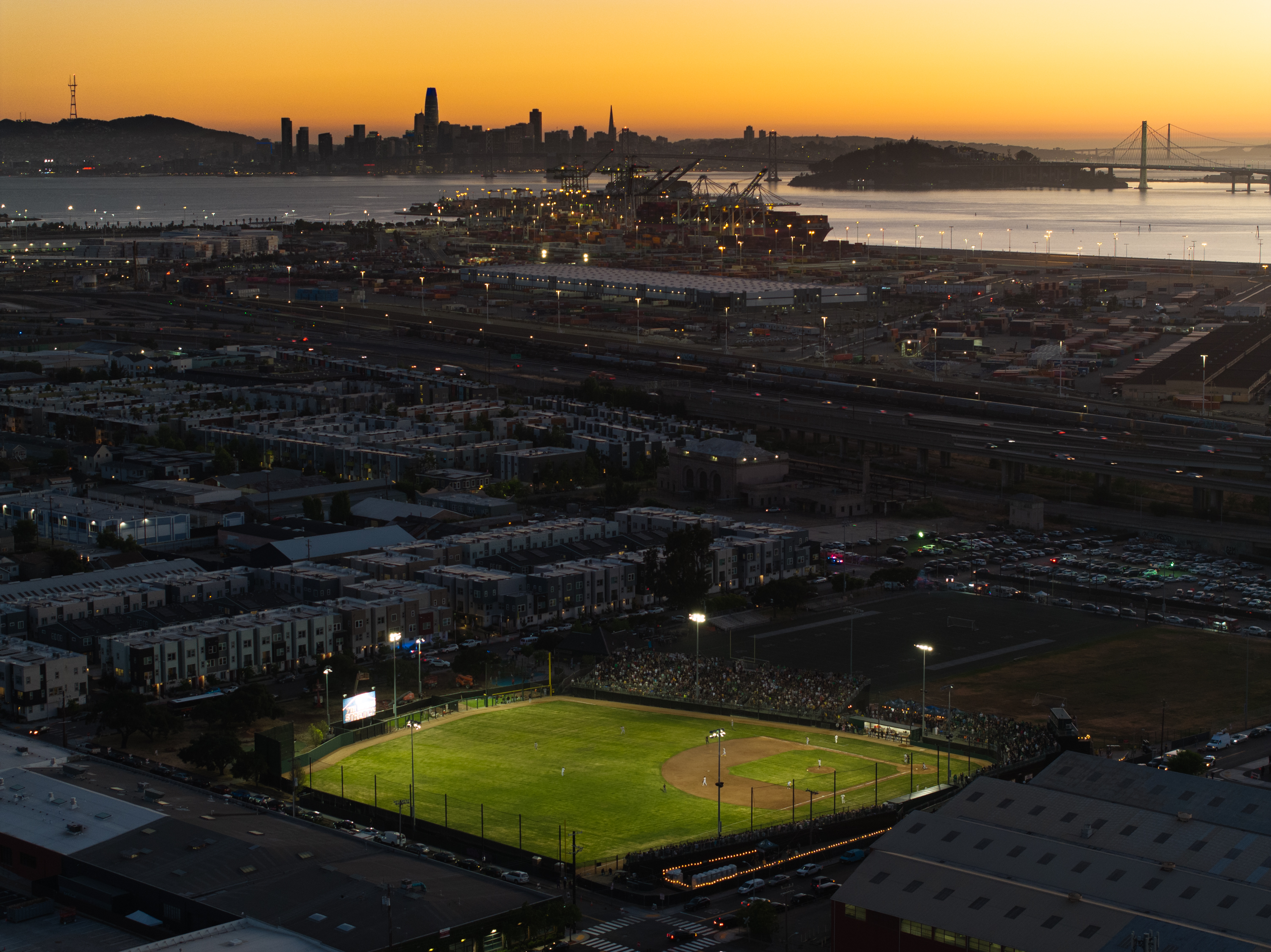 The image shows a baseball field lit up for a night game, surrounded by a cityscape, with a sunset over a water body and distant bridge and city skyline.