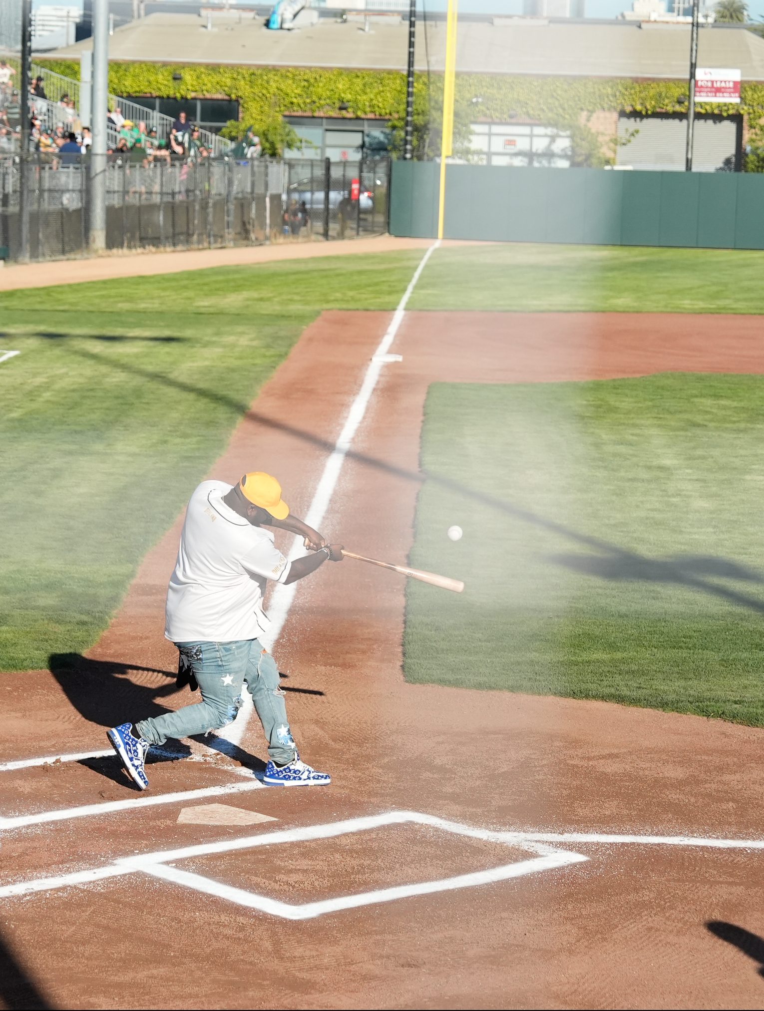 A person in a yellow cap and distressed jeans is swinging a bat at a baseball on a field, with the ball in mid-air and spectators visible in the background.