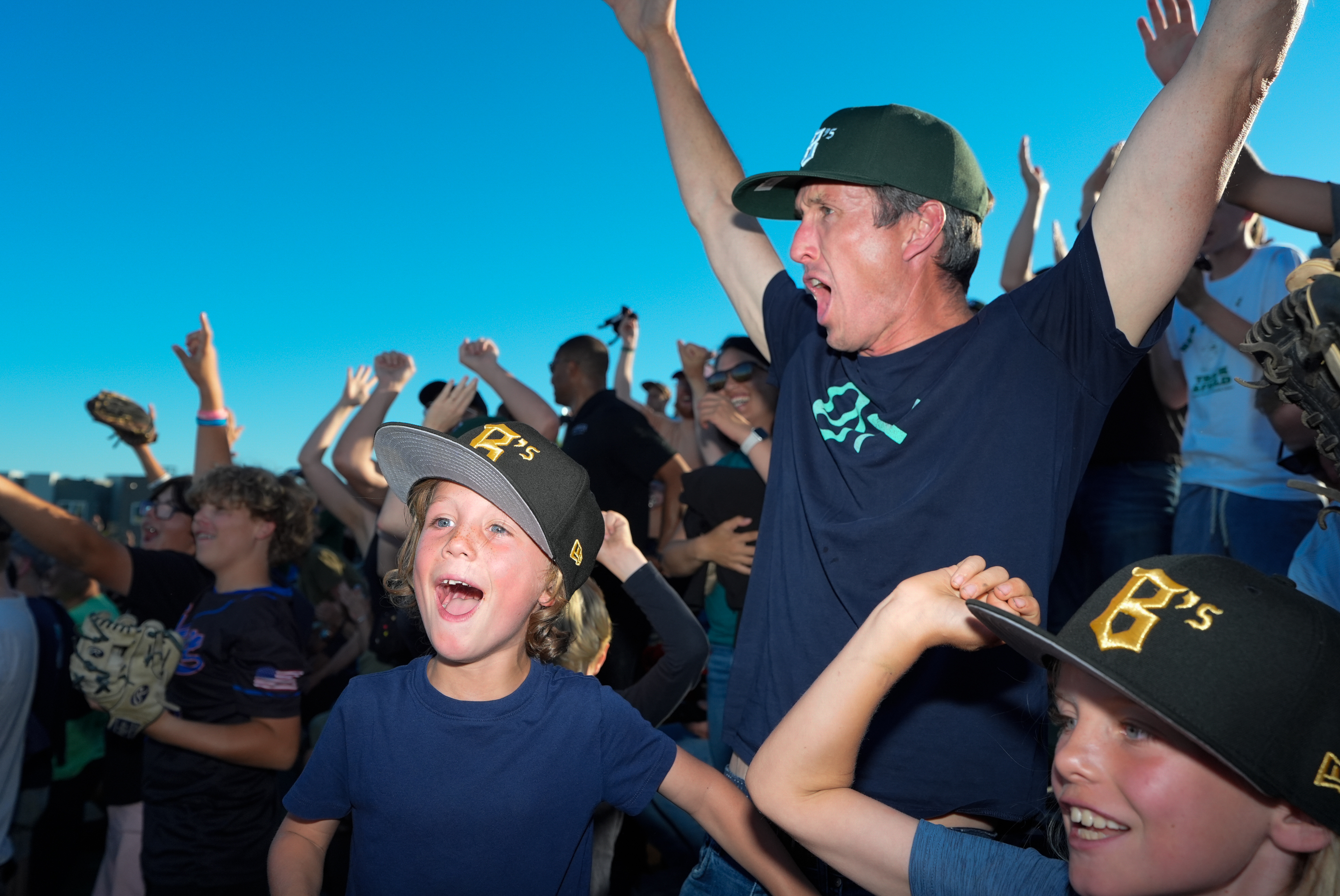 The image shows excited fans, including children and adults, cheering with their hands in the air. They are wearing matching team hats and appear to be at a sporting event.