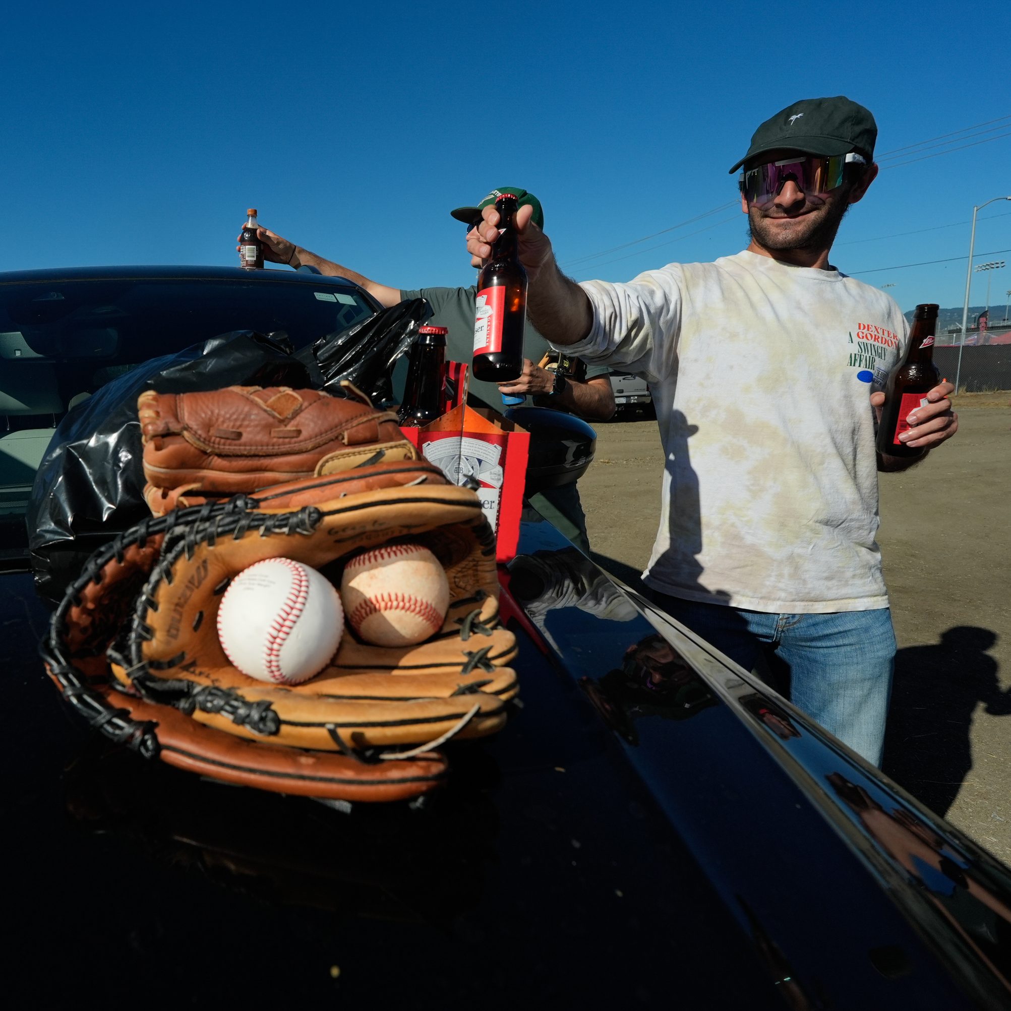 A person in casual clothes holds a beer bottle, standing by a car with baseball gloves, balls, and more bottles on the car's hood, under a clear blue sky.