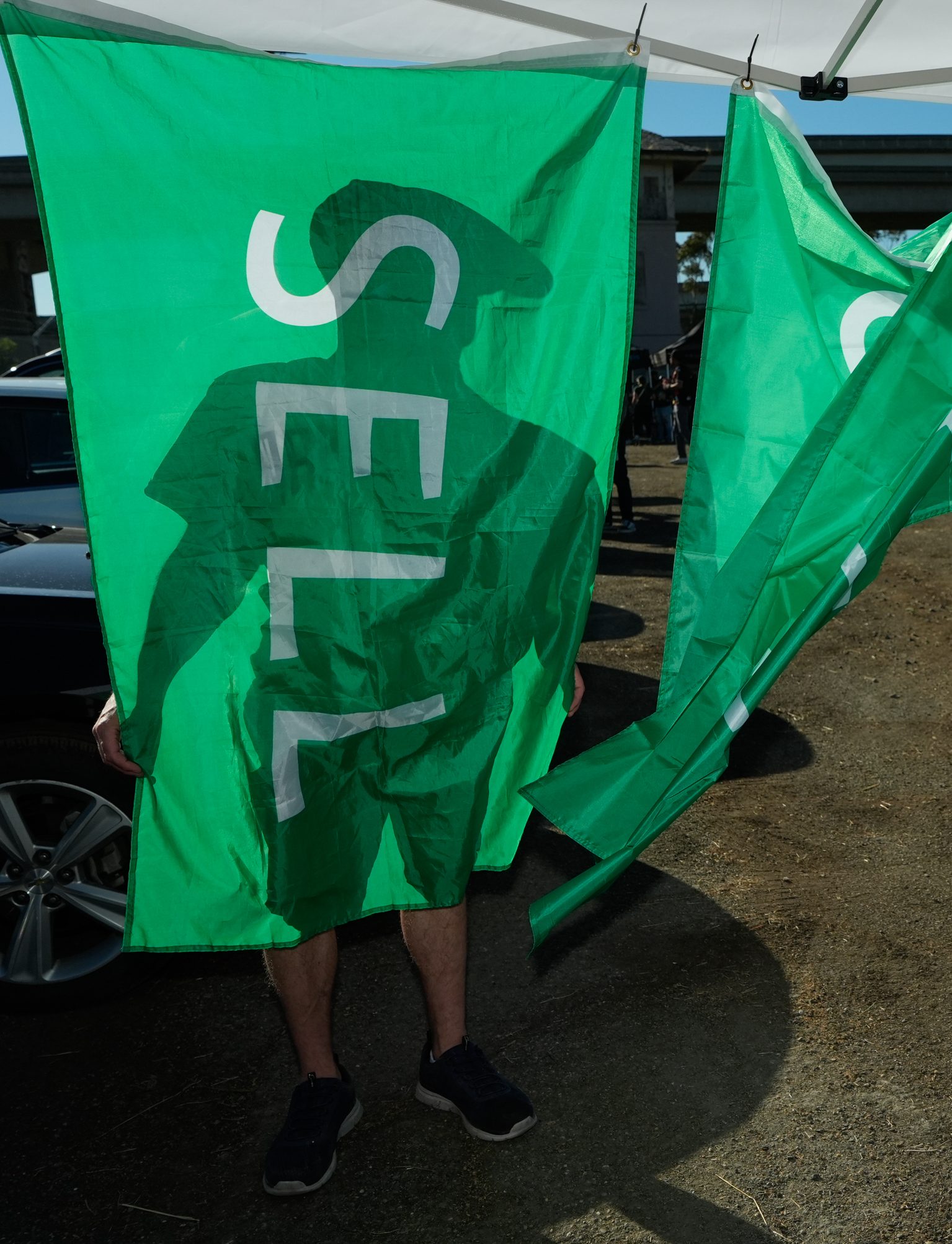 A person stands behind a green flag that reads &quot;SELL,&quot; casting a shadow. Only their legs and shoes are visible from beneath the flag.