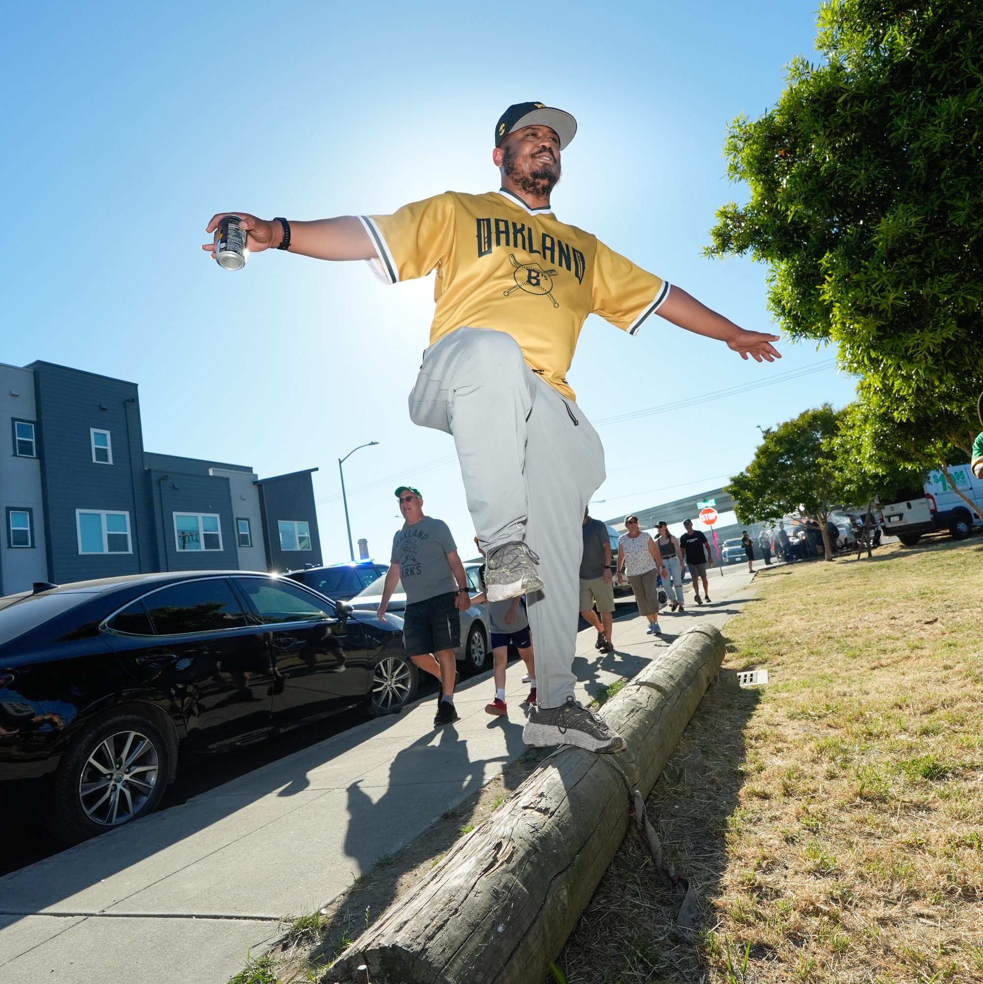 A man balances on a log while holding a drink. He is wearing an Oakland jersey and cap. People walk on the sidewalk next to parked cars in a sunny residential area.