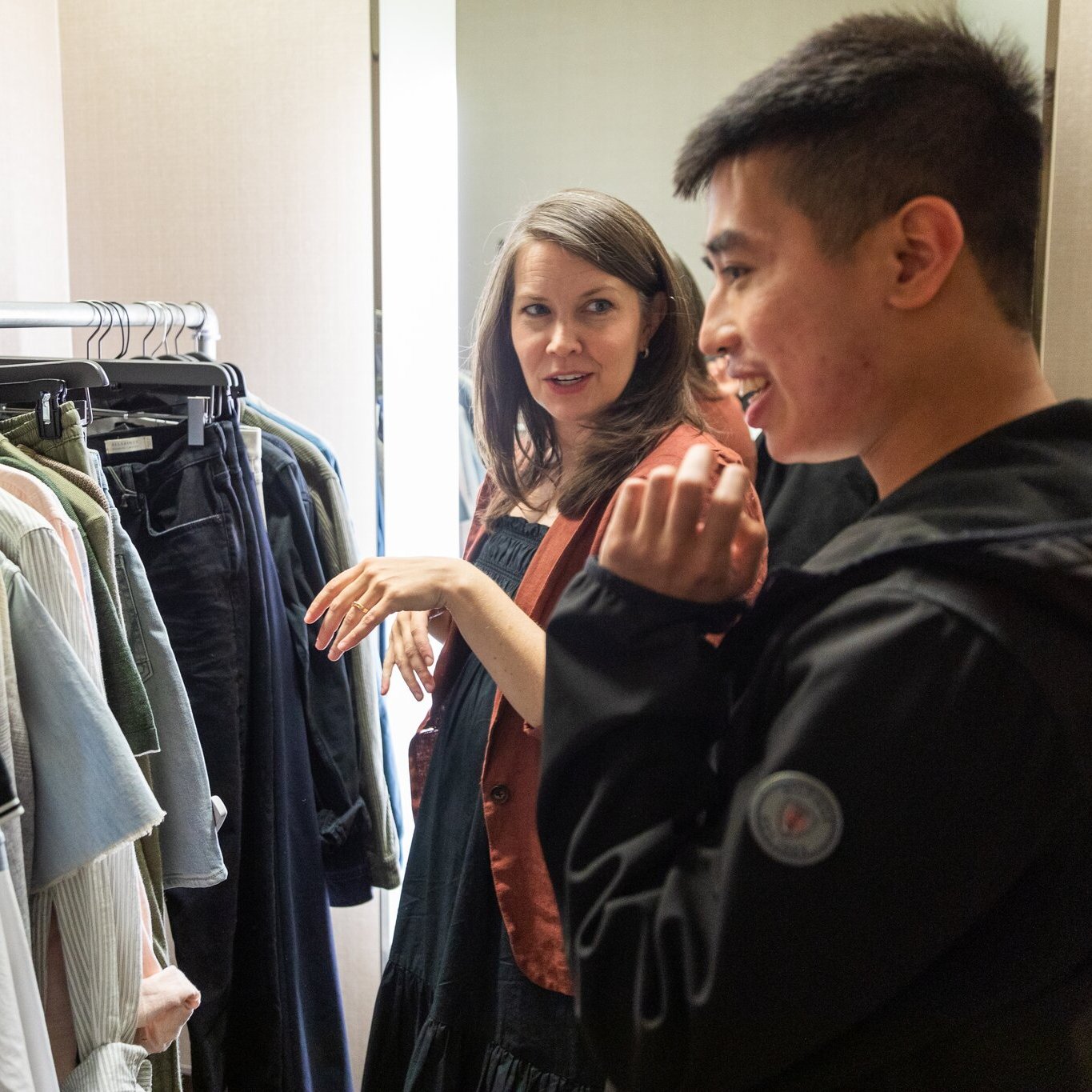 Two people are in a room with a clothing rack full of various garments. They are engaged in conversation, with one gesturing towards the clothes and the other smiling.