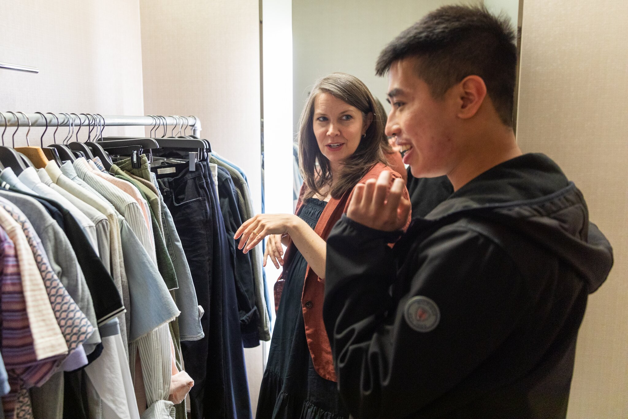 Two people are in a room with a clothing rack full of various garments. They are engaged in conversation, with one gesturing towards the clothes and the other smiling.