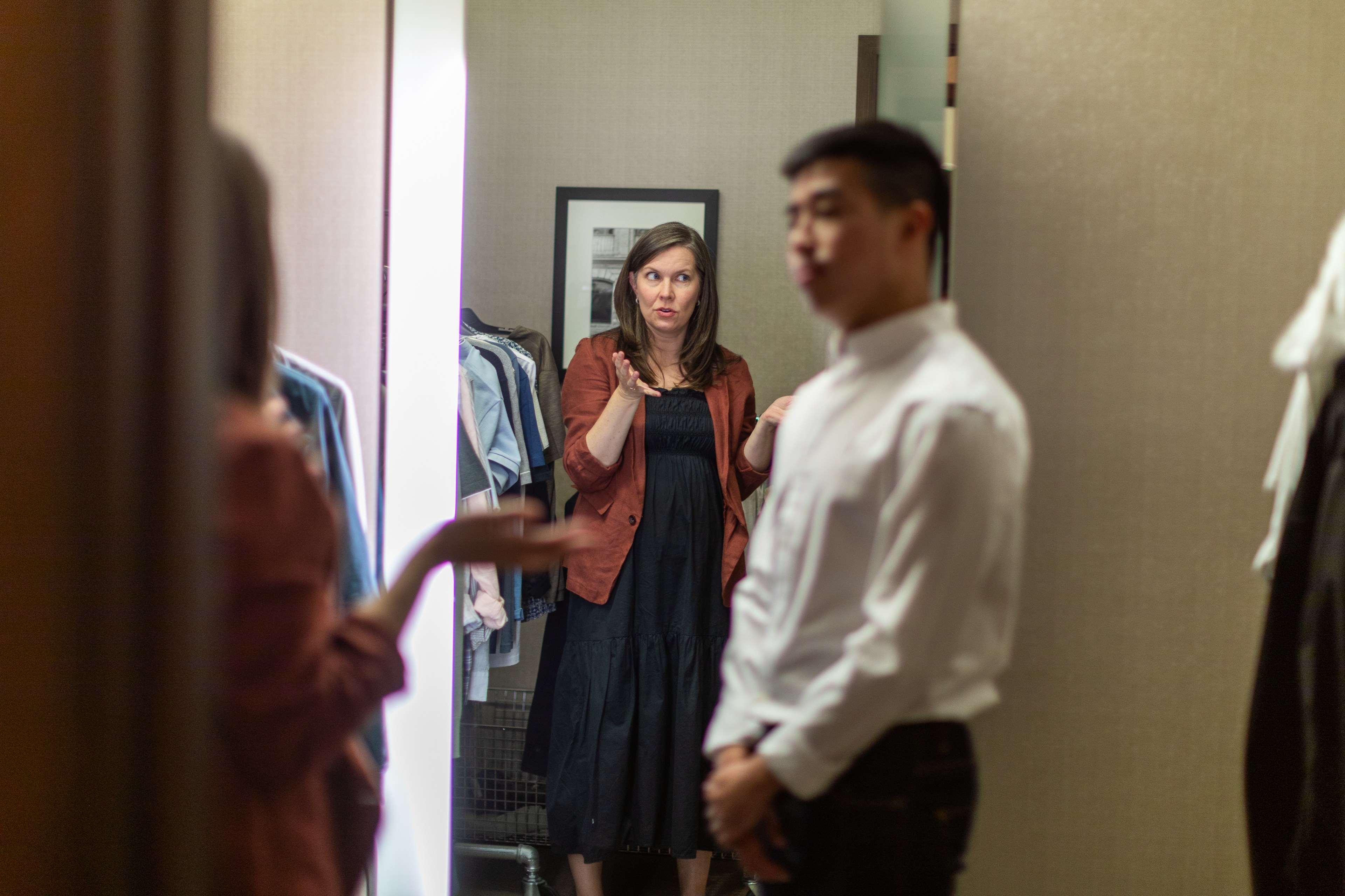 A woman in a brown jacket and black dress stands in front of a mirror, gesturing, while a man in a white shirt stands nearby. Clothing racks are visible.