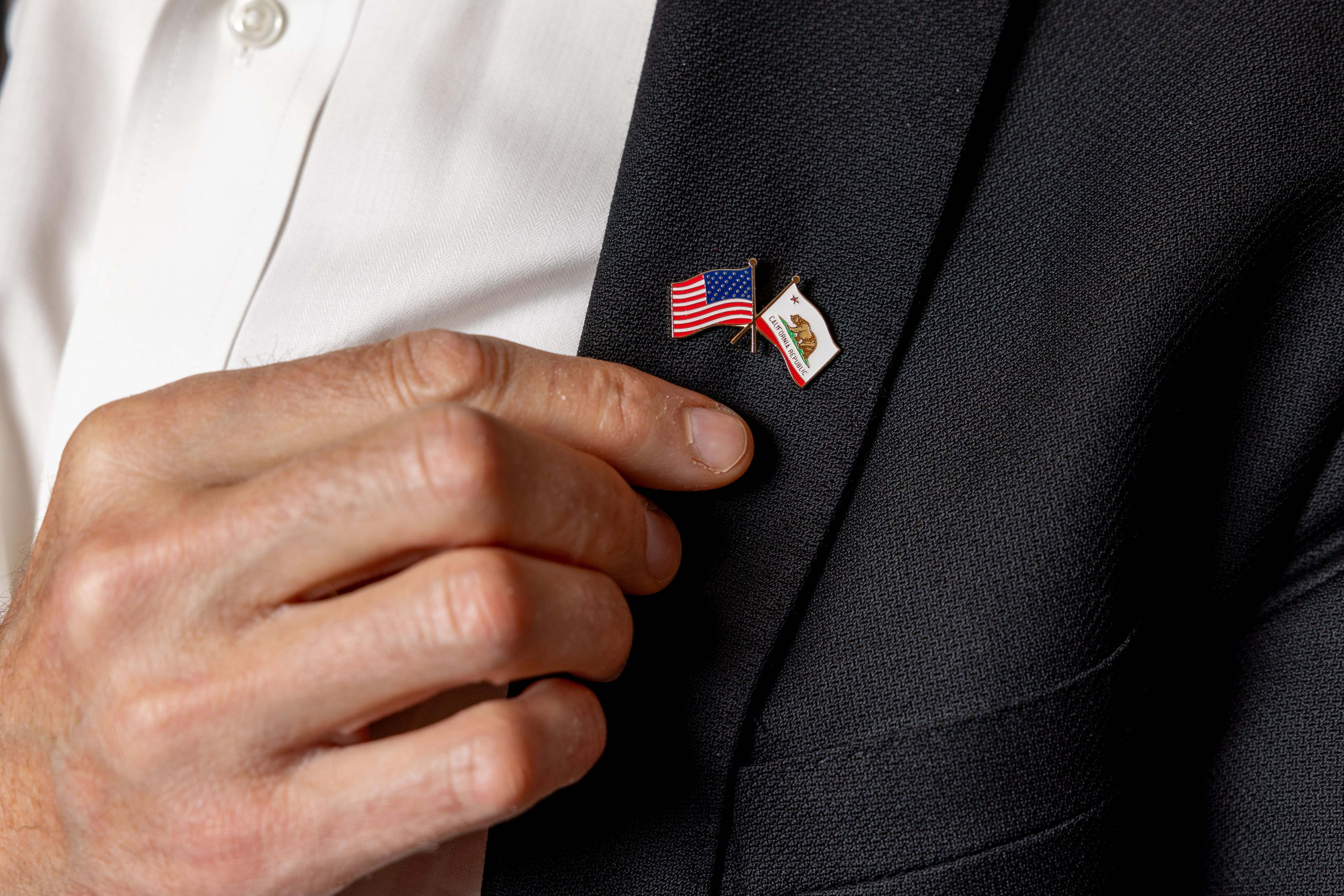 A hand is adjusting a lapel pin that features crossed American and Californian flags, attached to a dark-colored blazer worn over a white shirt.