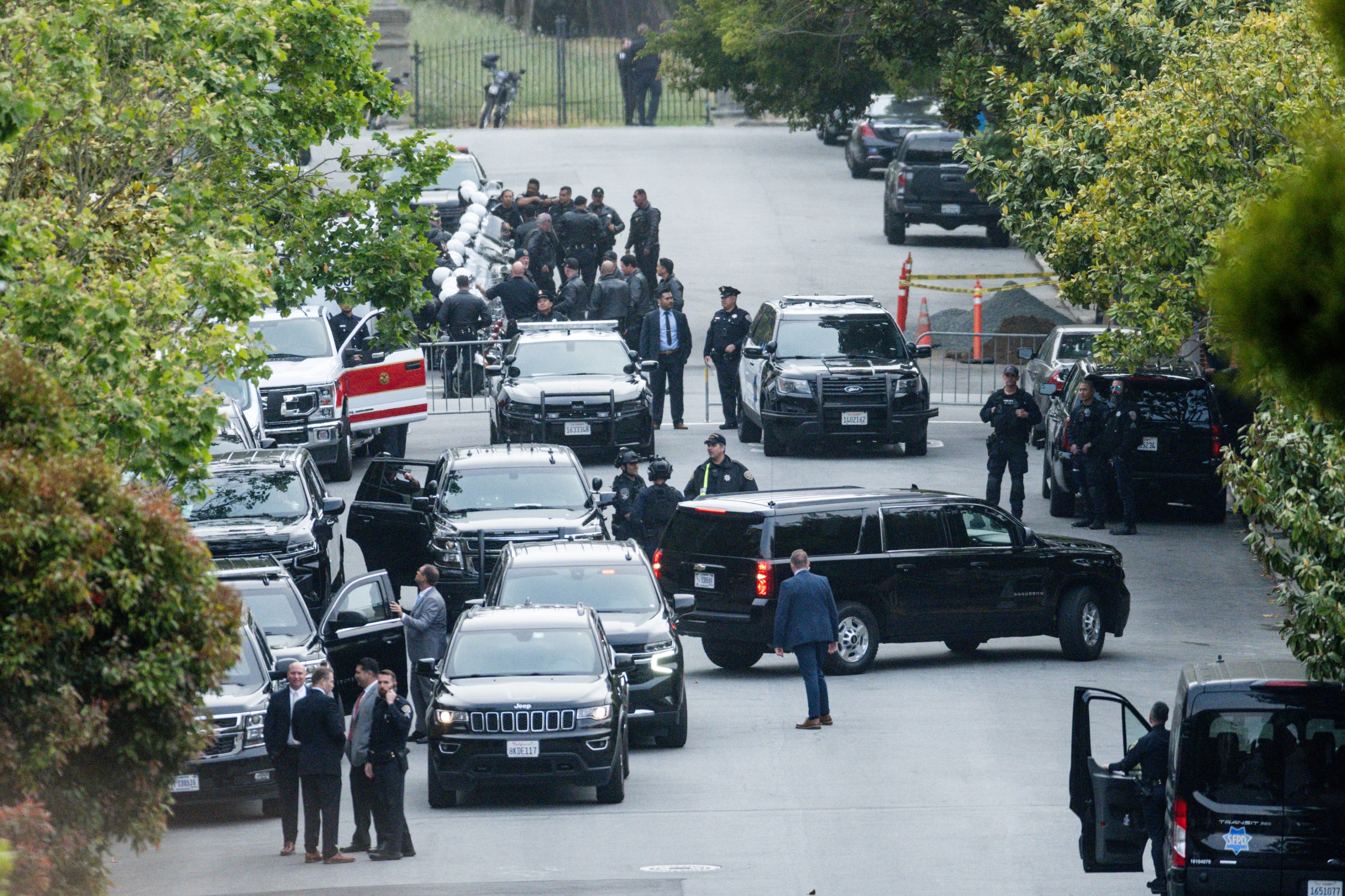 A large group of police officers and security personnel surround several black SUVs on a tree-lined street, creating a visible security detail.