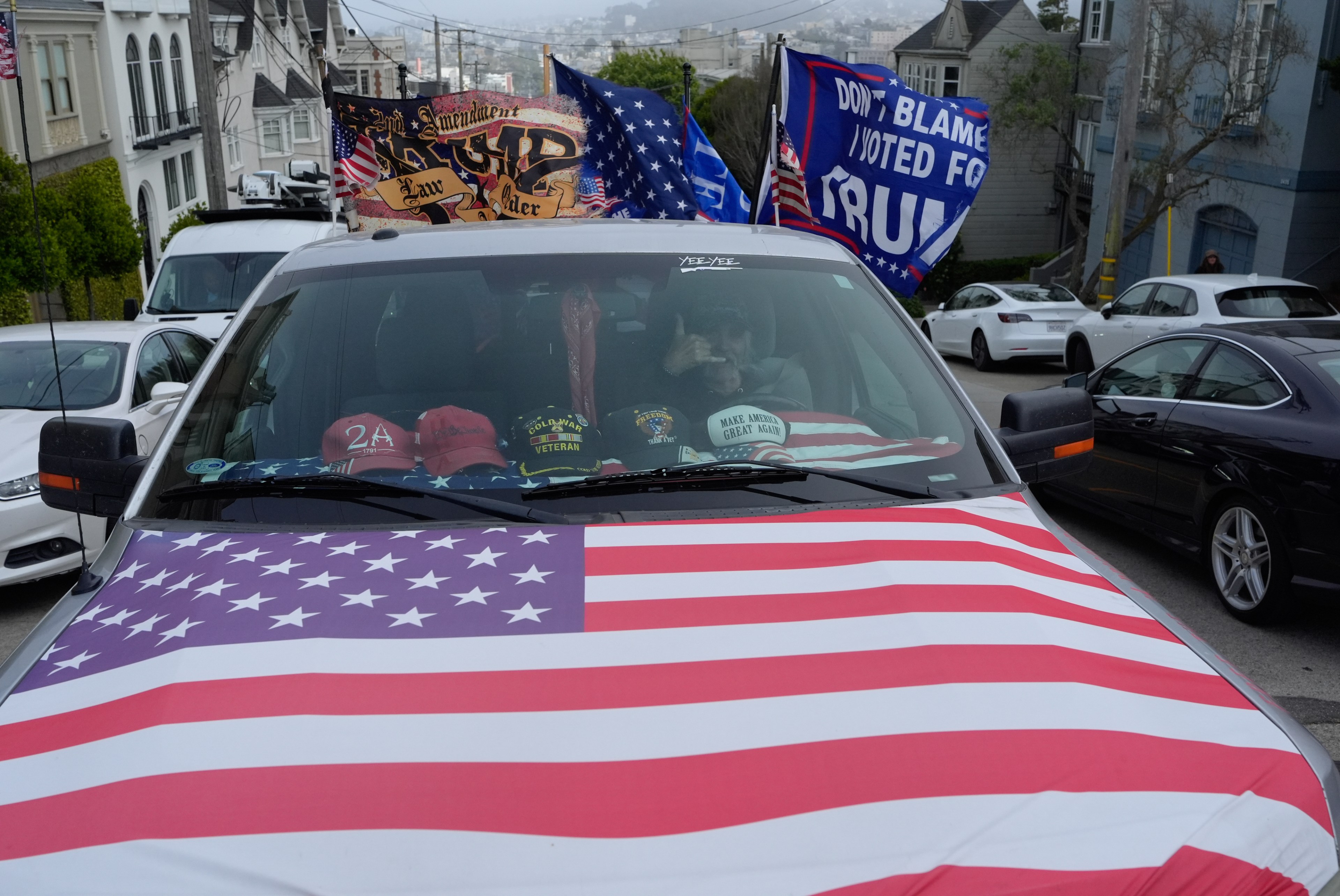 A truck draped in an American flag has its dashboard adorned with multiple hats, and pro-Trump flags are attached. It is parked on a street with other cars.