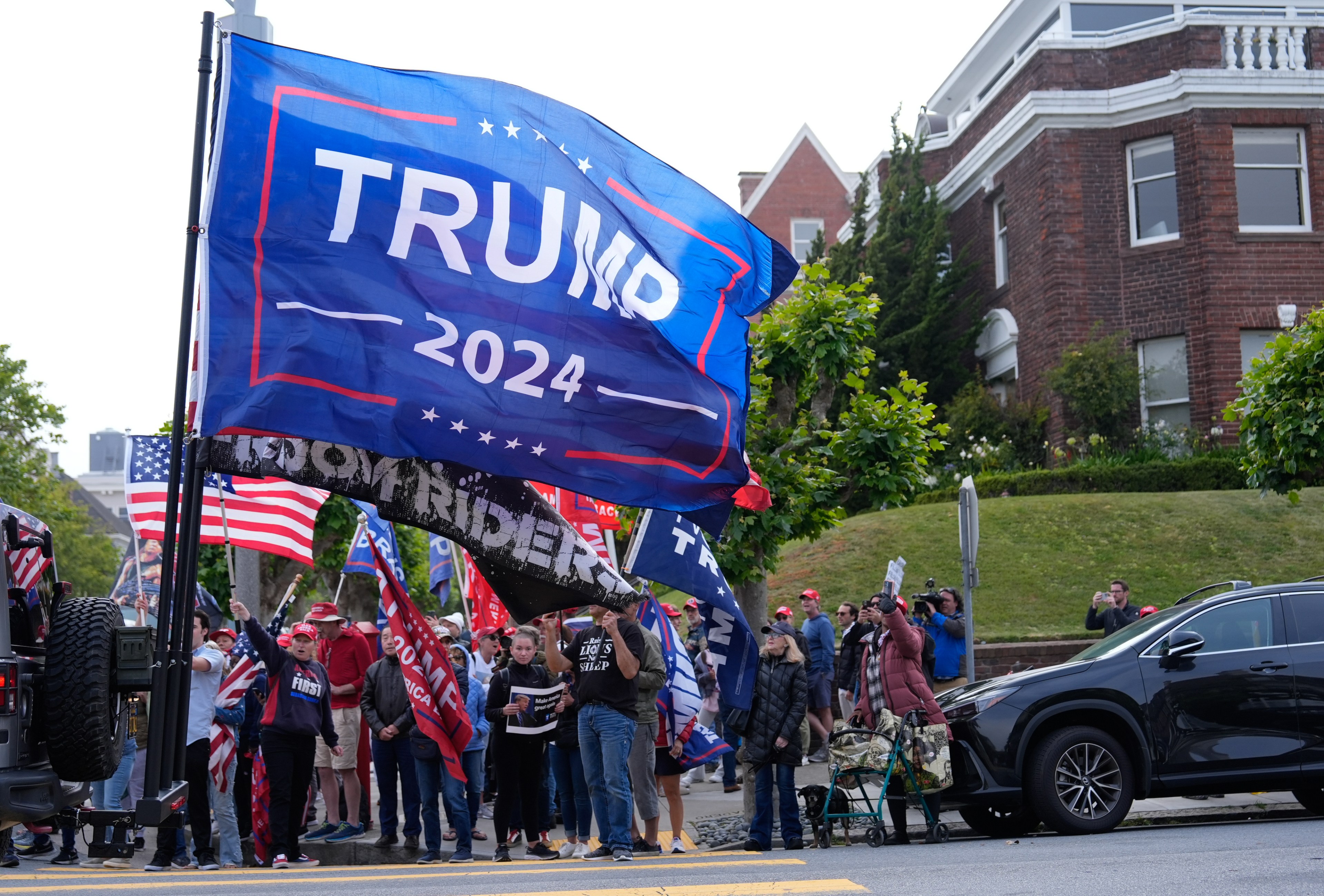 A group of people is gathered on a street, holding flags and signs supporting Trump for the 2024 presidential campaign. A large banner reads &quot;TRUMP 2024.&quot;