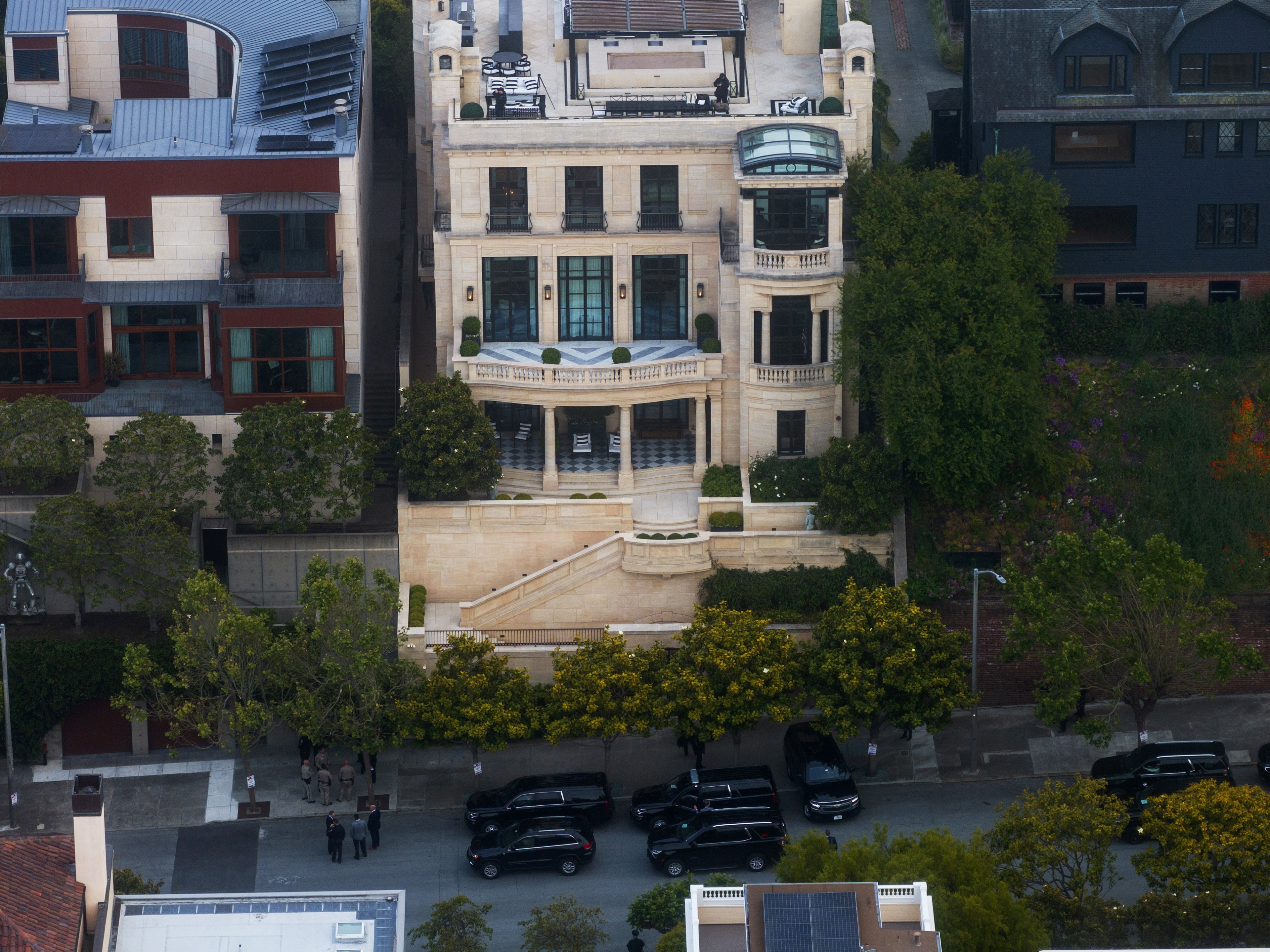 An aerial view of a grand beige mansion with a rooftop terrace, lush greenery, and several black cars parked on a tree-lined street below.