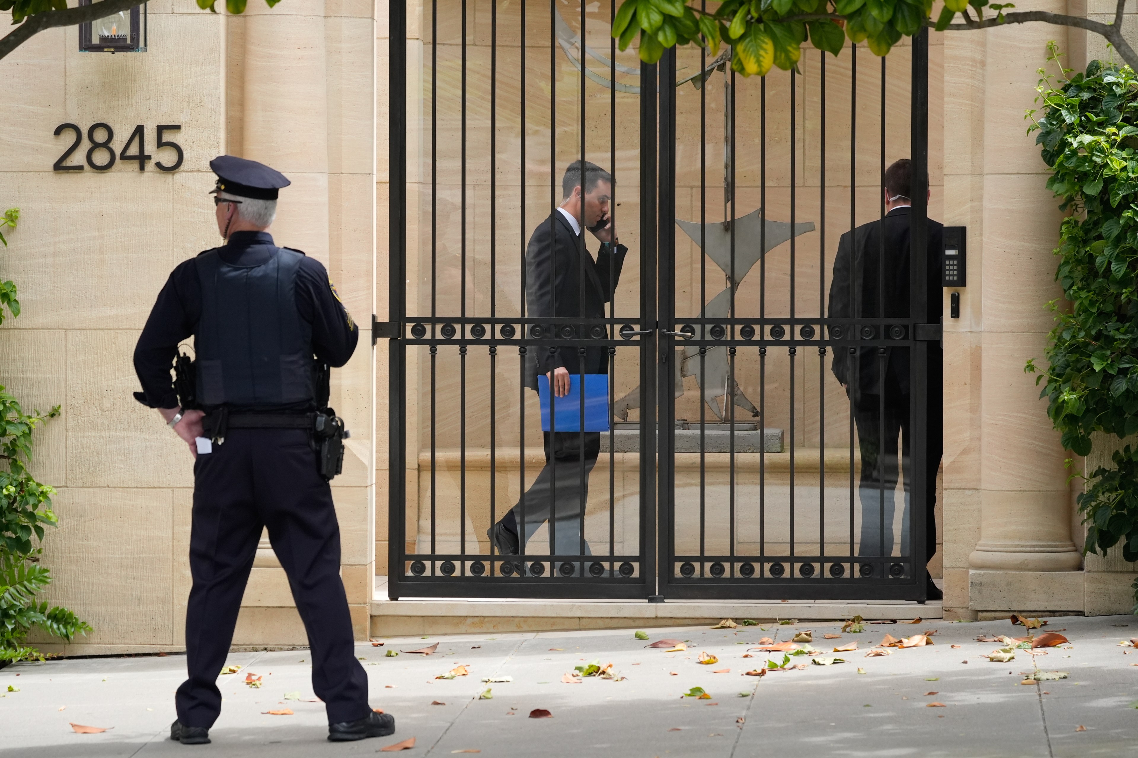 A police officer stands guard outside a gated building at 2845, while two men in suits enter, one using a keypad and the other on the phone, holding a blue folder.