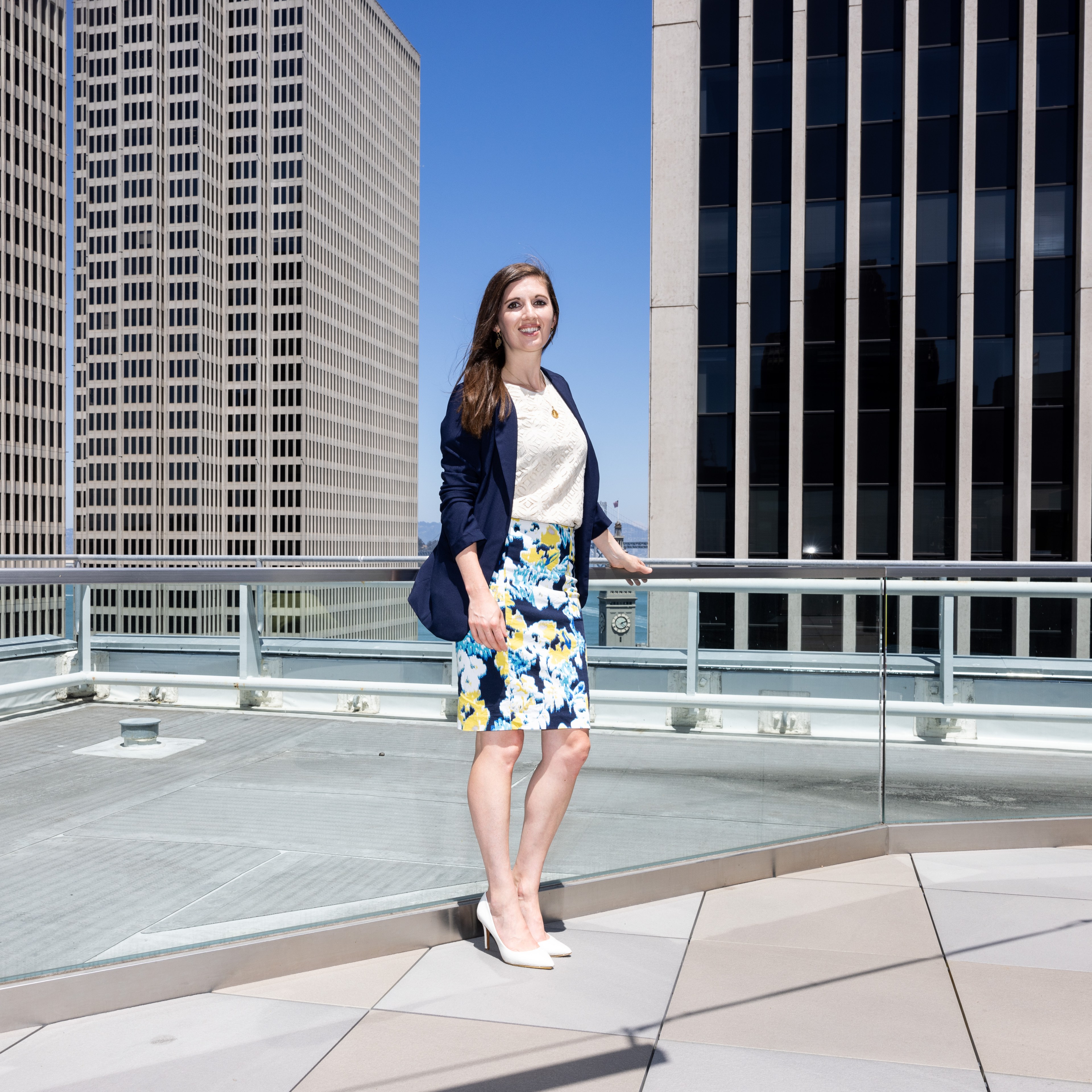 A woman stands on a rooftop, surrounded by tall buildings. She is dressed in a navy blazer, white blouse, a floral skirt, and white heels, smiling at the camera.