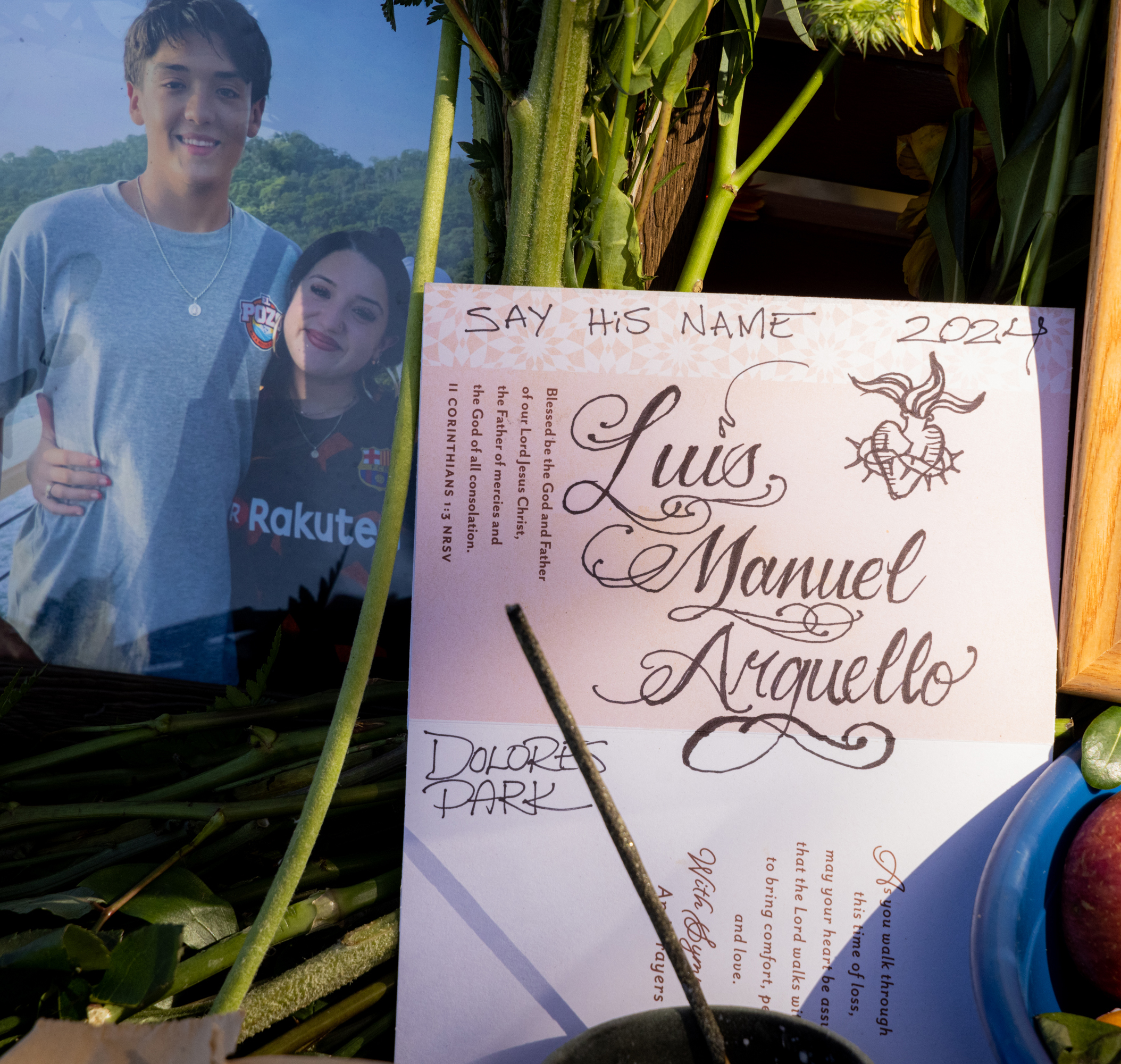 The image shows a remembrance setup with a photo of two smiling young individuals and a memorial card for Luis Manuel Arguello, surrounded by flowers and an apple.