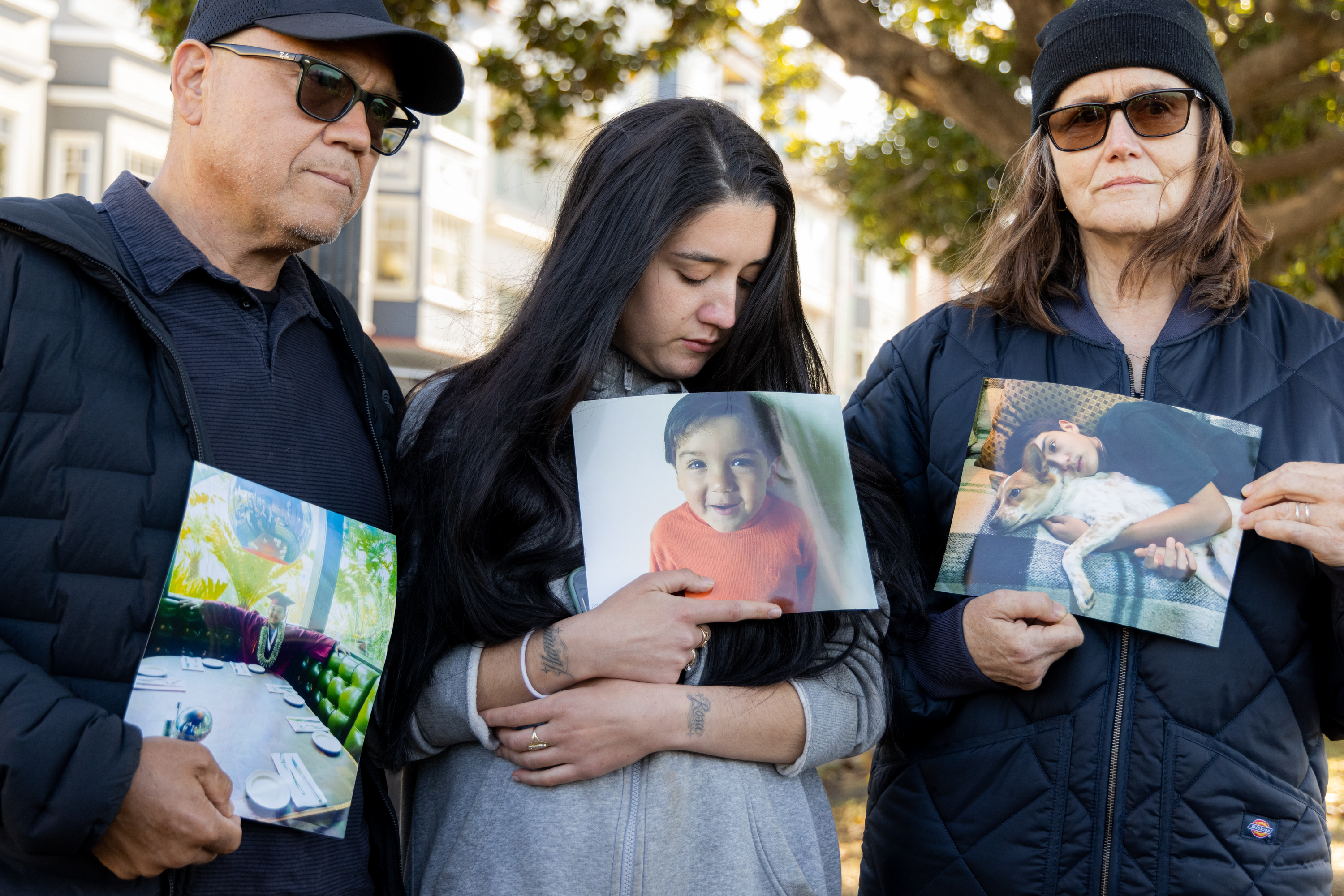 Three people stand holding photos, each looking solemn. The man on the left holds a picture of a colorful diner, the woman in the middle holds a child's photo, and the woman on the right holds a boy with a dog.