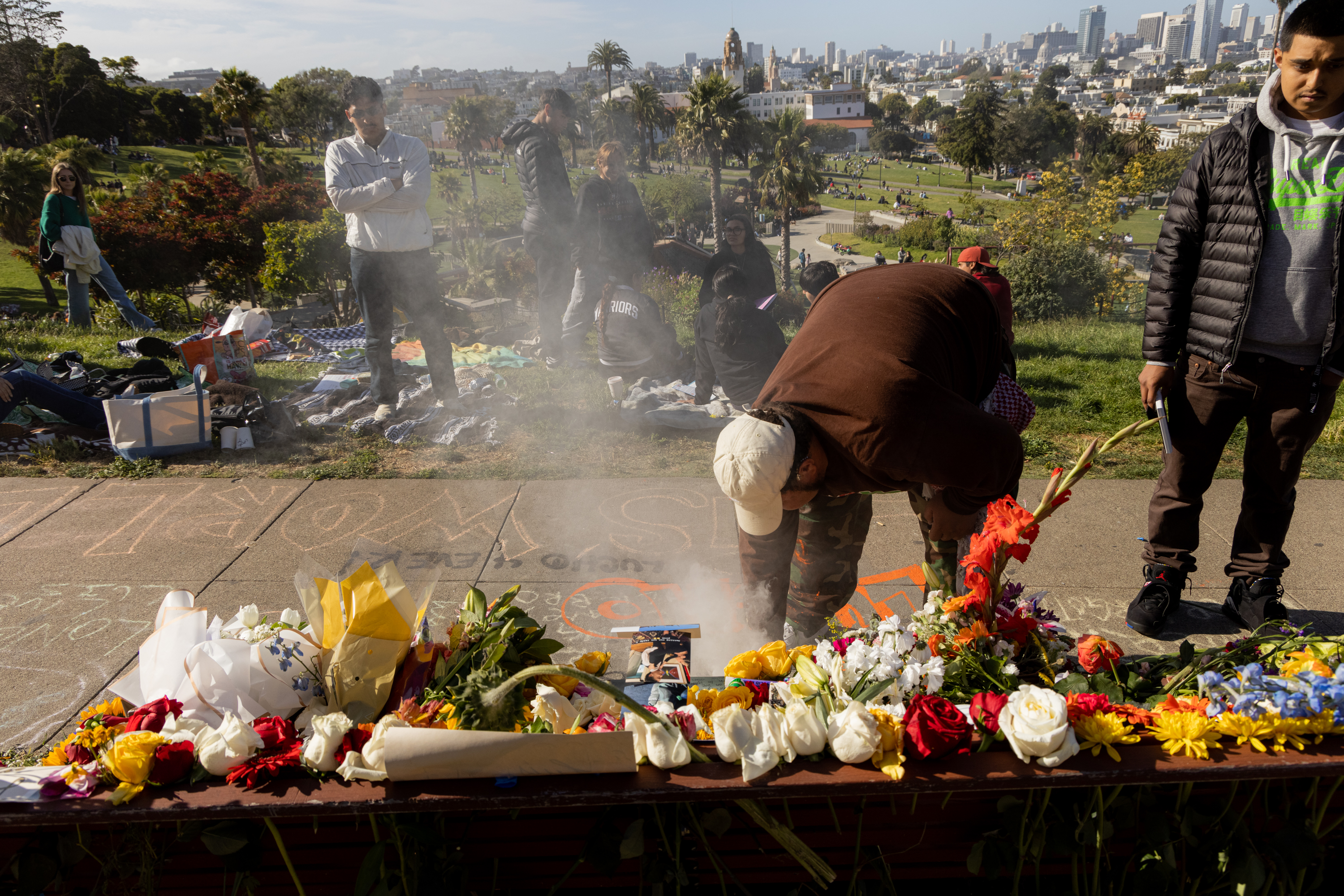In a park, people gather near vibrant flowers and a memorial where a person bends over, creating smoke, while others stand or sit on blankets around the area.