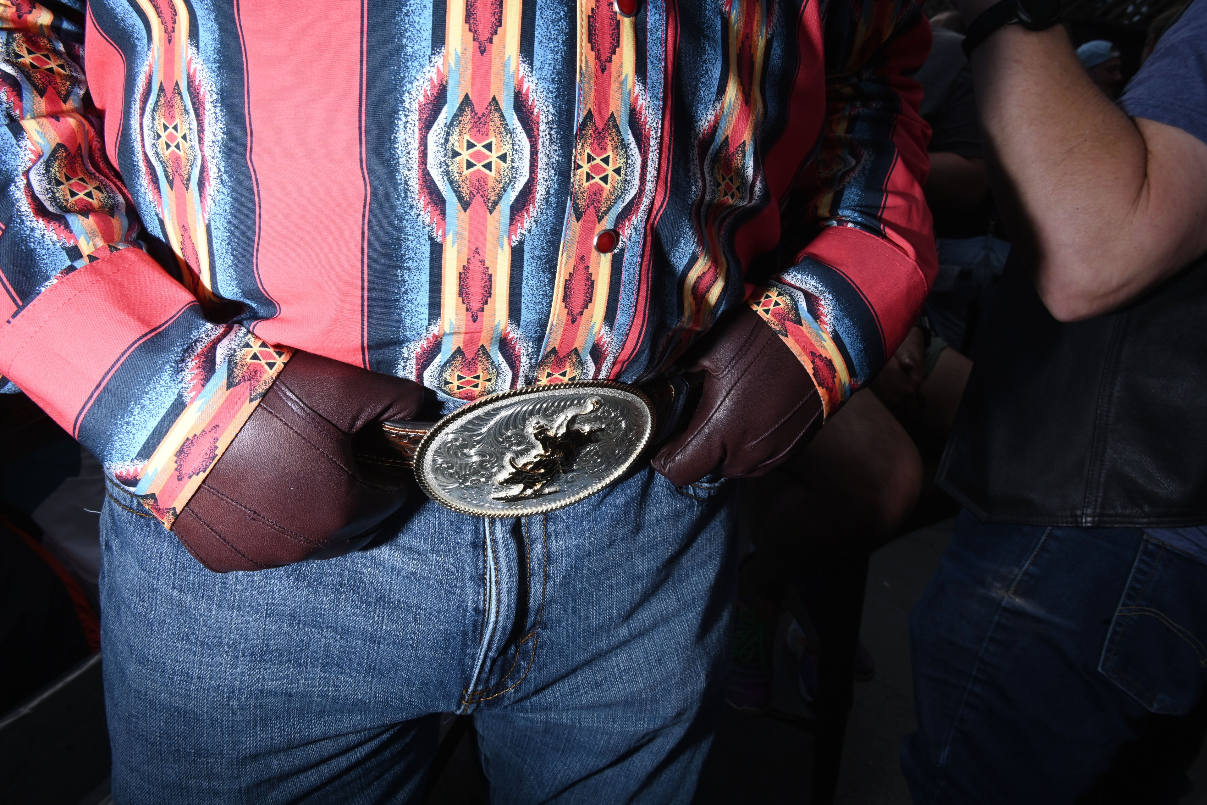 A person wears a colorful patterned shirt, blue jeans, and brown leather gloves, with hands resting on a large ornate silver belt buckle.