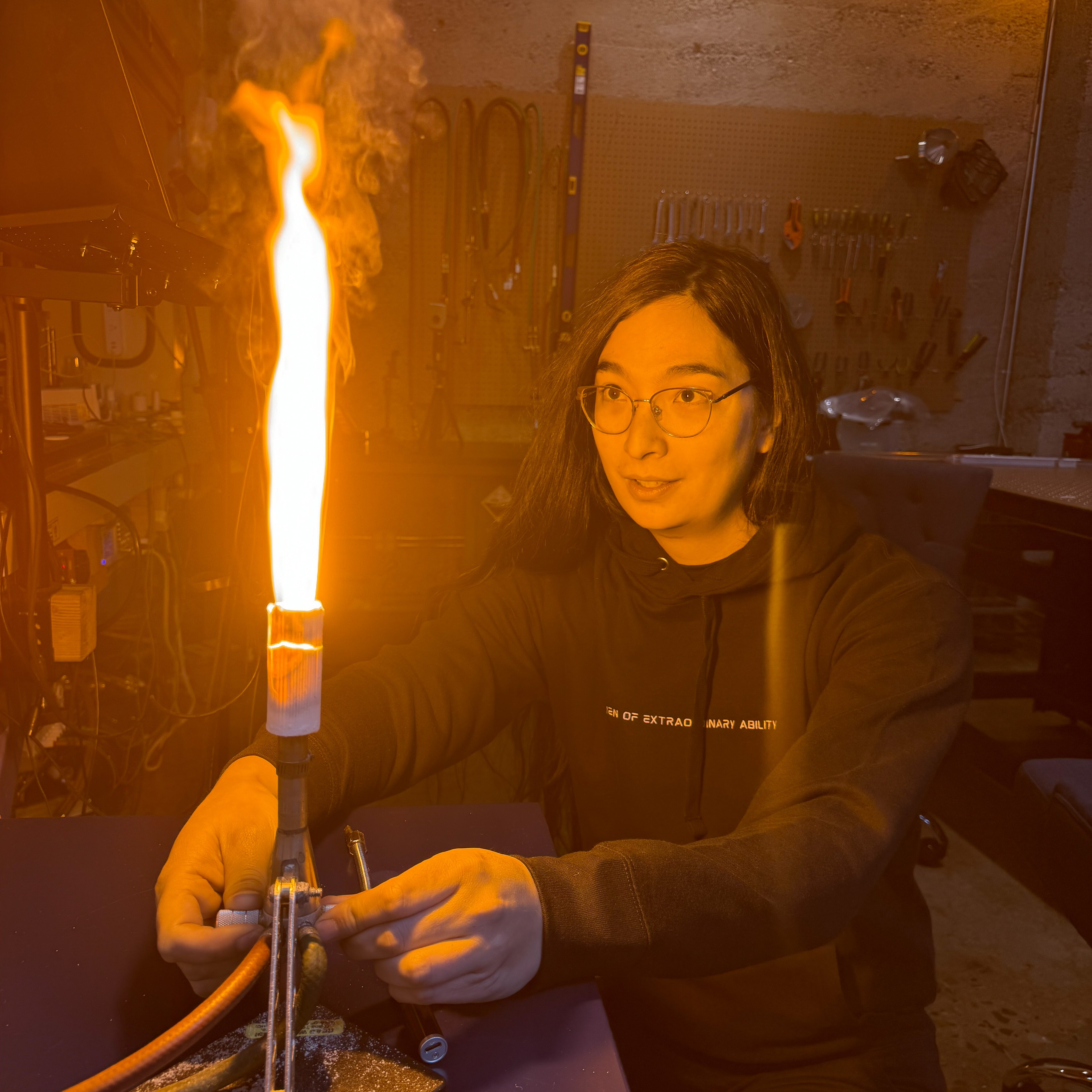 A person in glasses and a black hoodie adjusts a setup with a large, bright flame, appearing to conduct an experiment in a workshop with tools hanging on the wall.