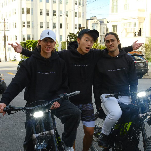Three friends wearing black hoodies pose on electric bikes; two with glasses and one in a white cap. They stand together on a sunny urban street with buildings behind them.