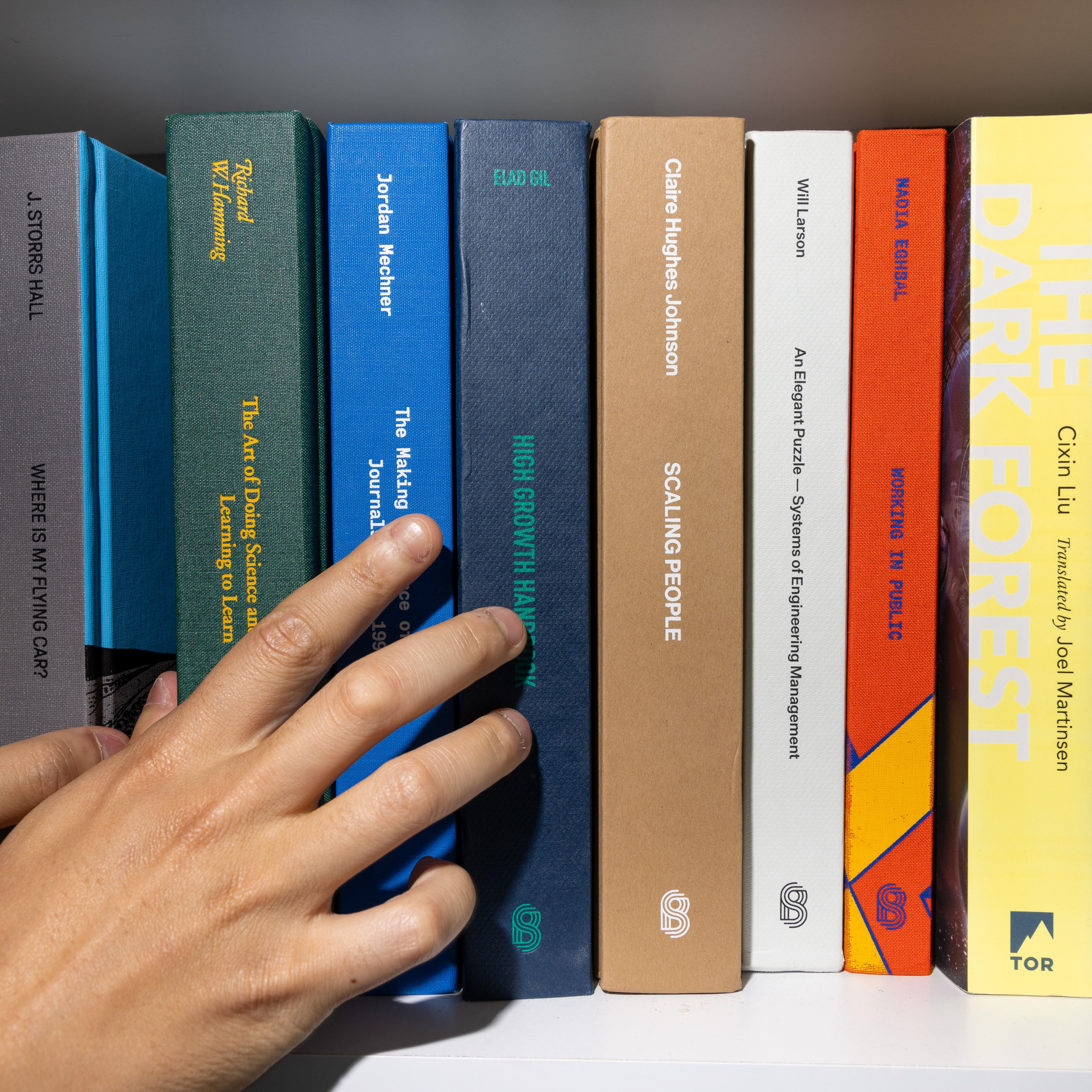 A hand is selecting a book from a neatly arranged bookshelf that features titles such as &quot;Scaling People,&quot; &quot;The Dark Forest,&quot; and &quot;High Growth Handbook.&quot;