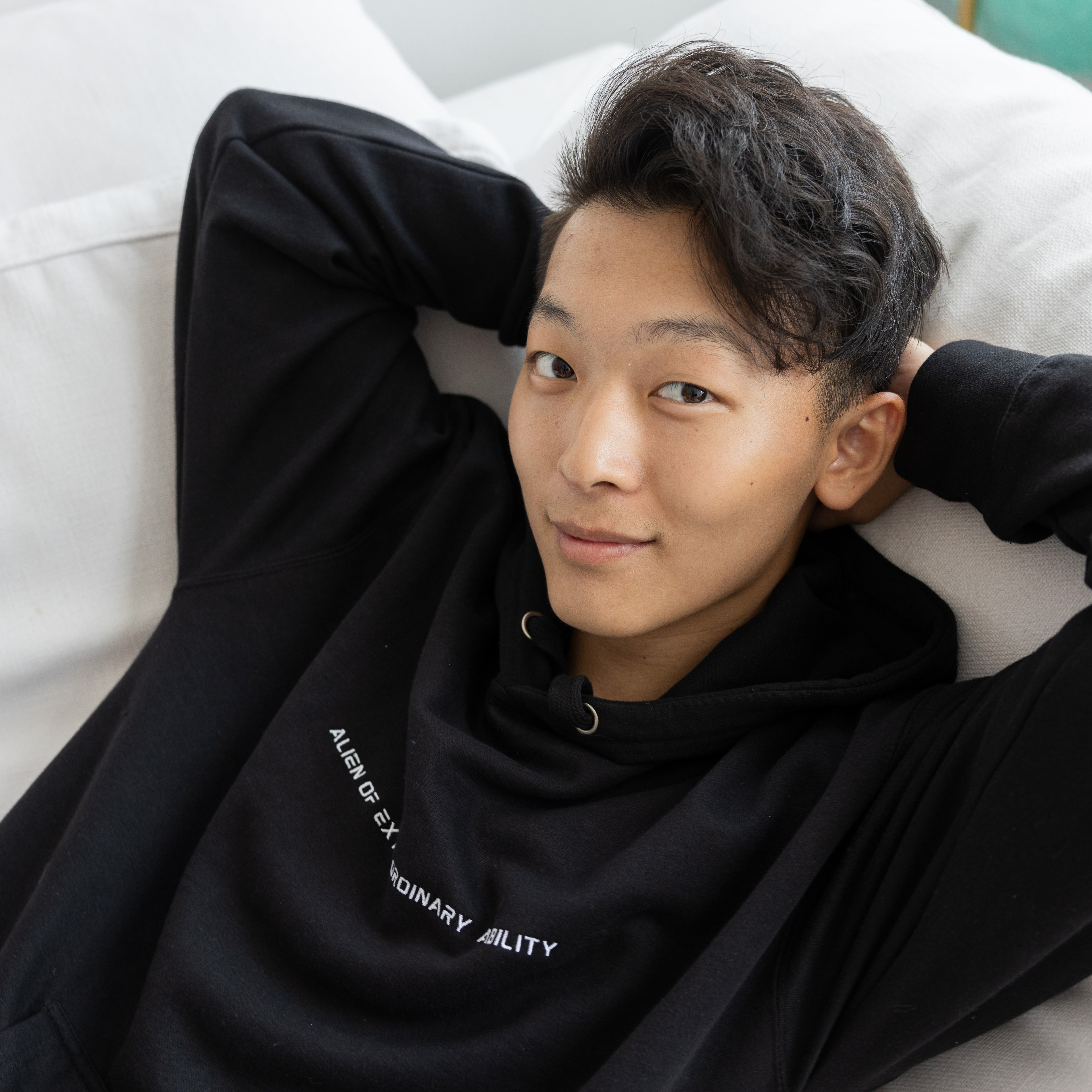 A young person with short hair, wearing a black hoodie with text on it, is lounging on a white couch, smiling with hands casually behind their head.