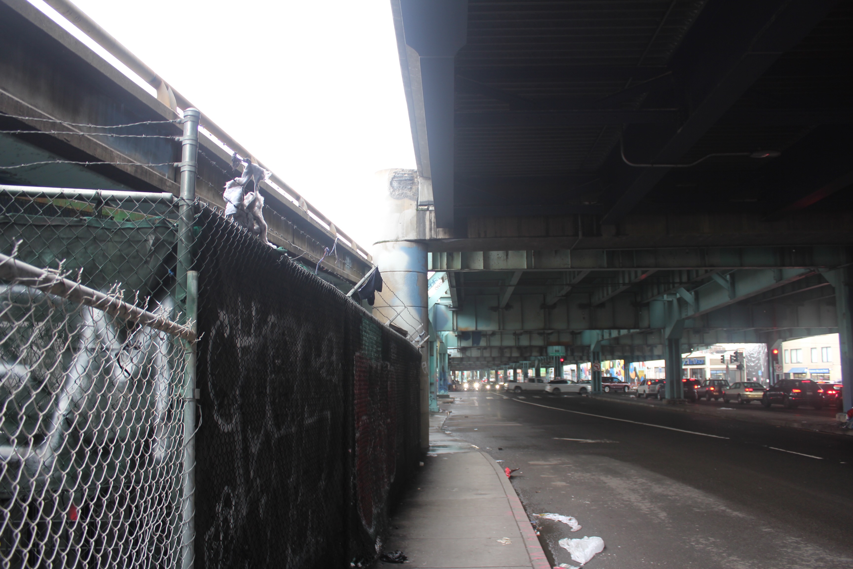The image shows a dimly lit, slightly wet street beneath an overpass, with a barbed-wire fence to the left, and light traffic in the background.