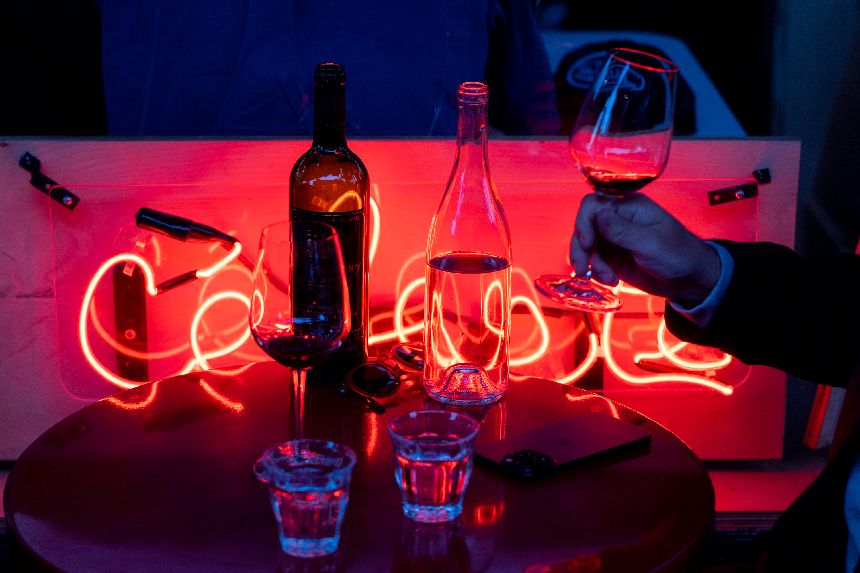 A hand holding a glass of red wine is visible, with a bottle, water glass, and phone on a table. A red neon light in the background creates a vibrant ambiance.