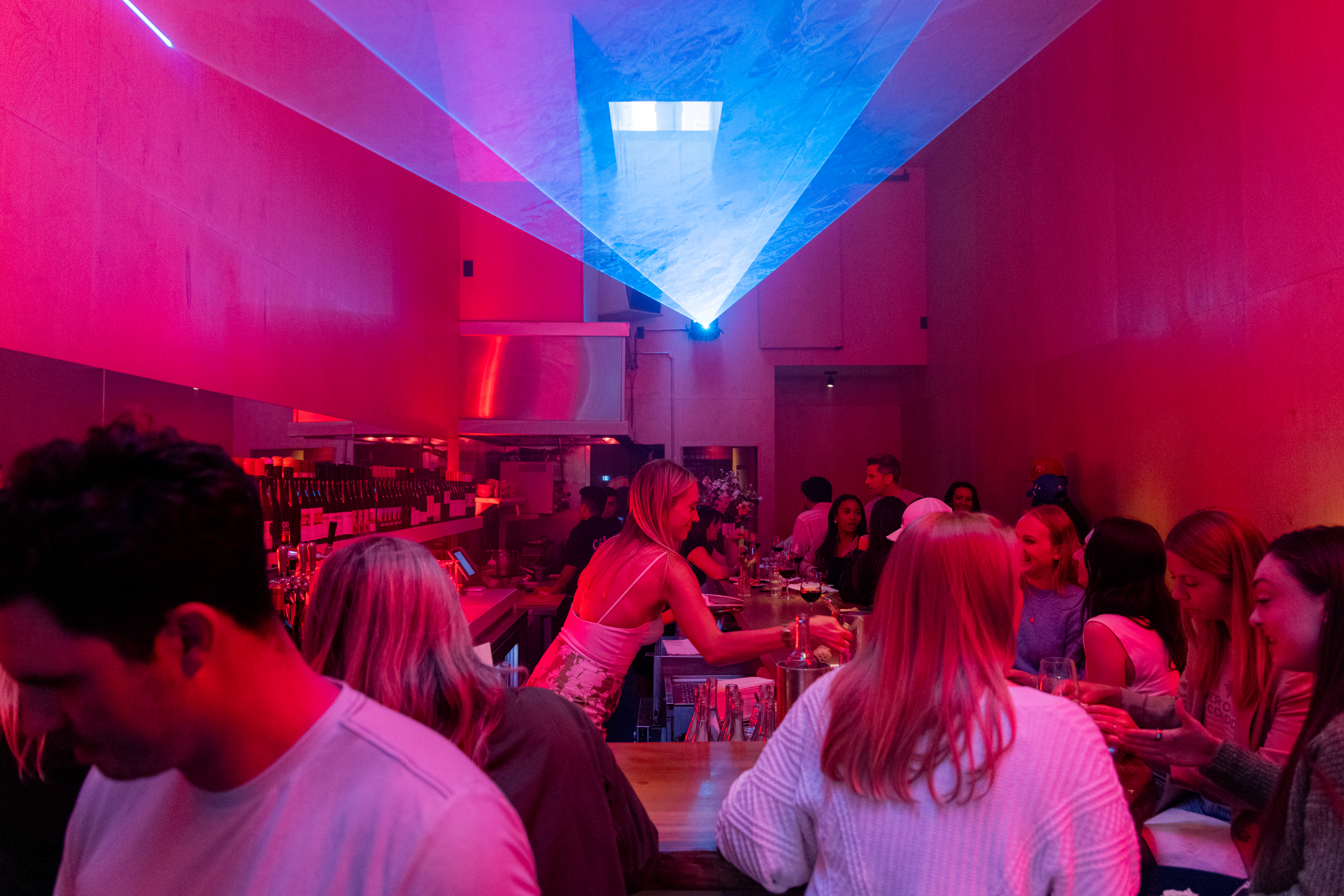 The image shows a vibrant bar scene illuminated by pink and blue lights. Patrons are socializing at the bar and surrounding tables, with drinks in hand. A bartender is seen interacting with customers.