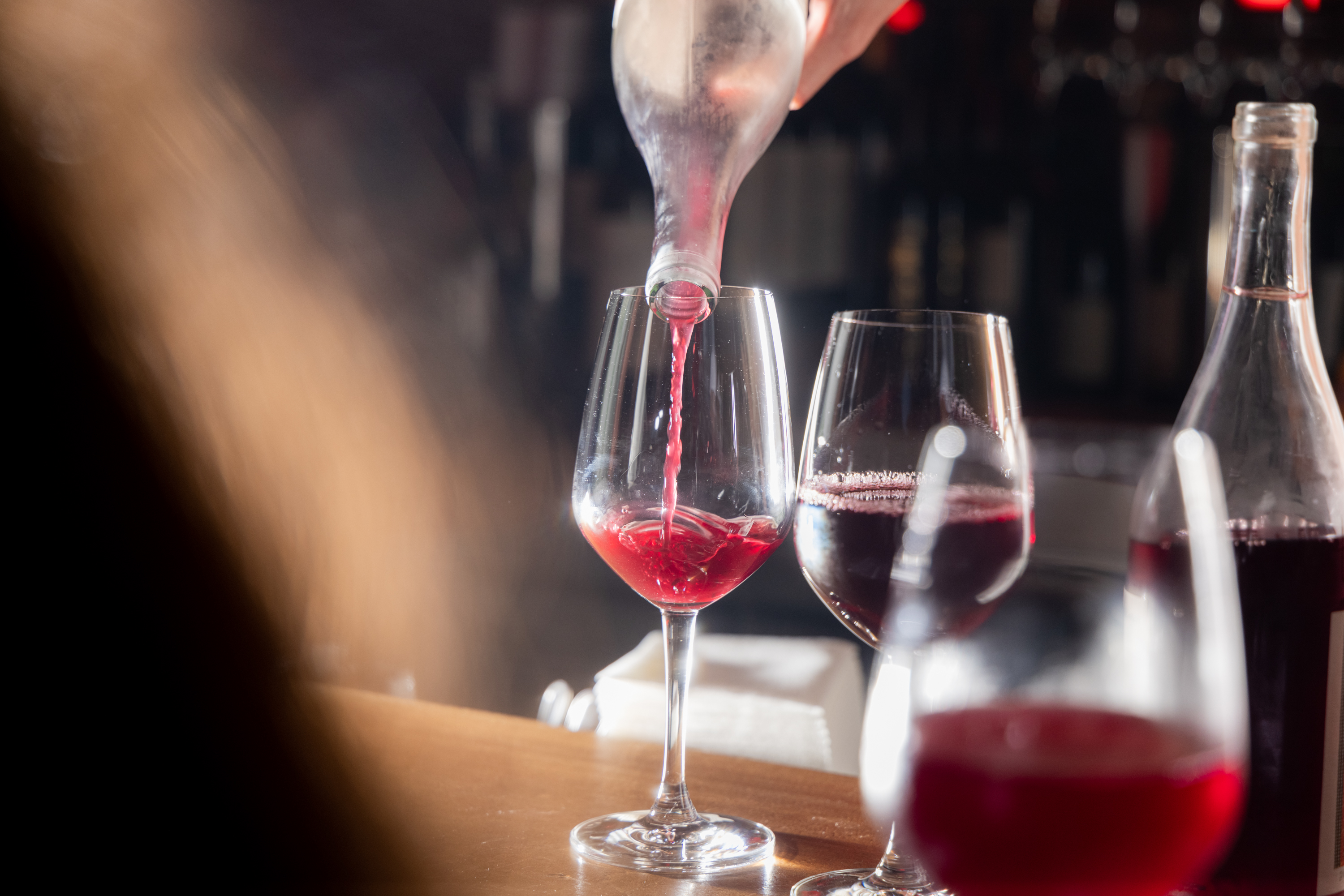 A hand is pouring red wine from a clear bottle into a wine glass on a bar counter, surrounded by other filled and partially filled wine glasses.
