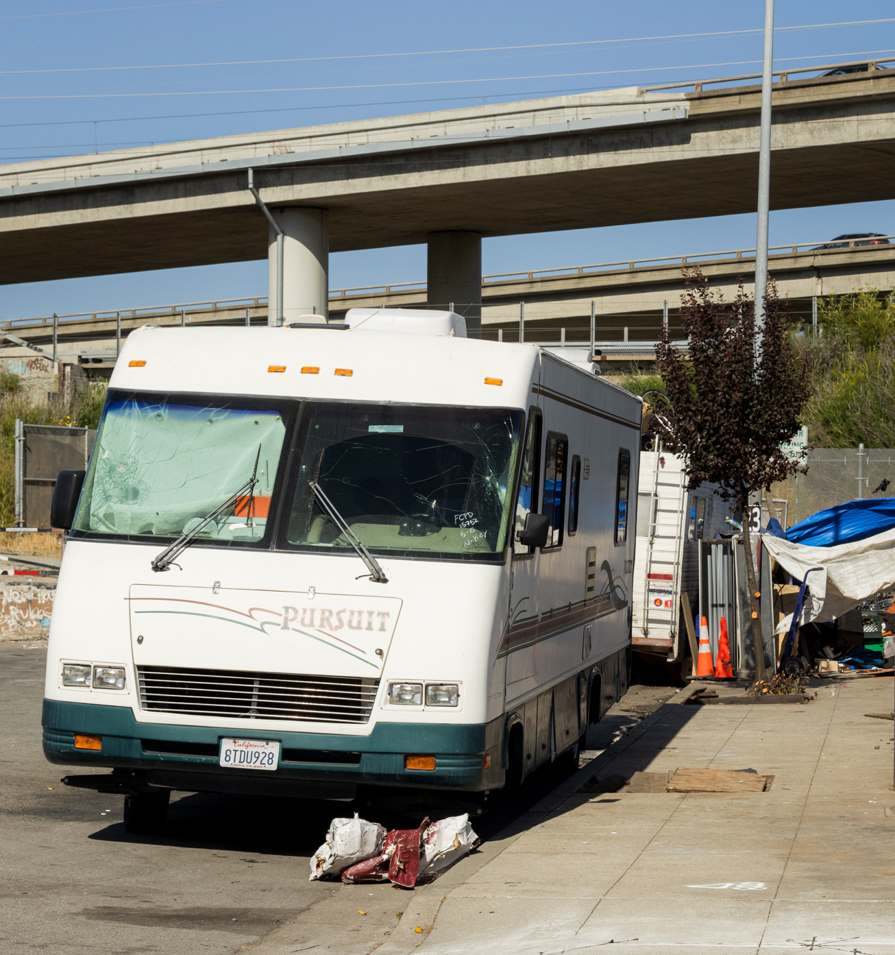 A white RV with &quot;Pursuit&quot; written on it is parked on the street near an overpass, surrounded by some debris and makeshift shelter materials.
