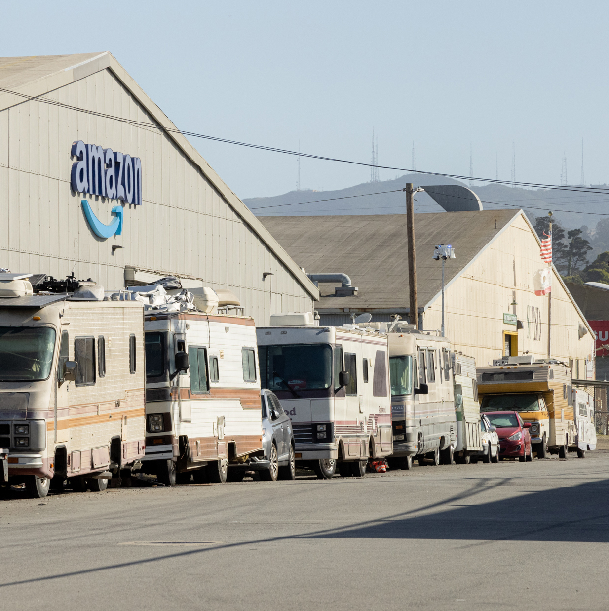 A row of RVs and cars are parked alongside a large building with the &quot;Amazon&quot; logo, backed by a mountainous landscape and electrical towers in the distance.