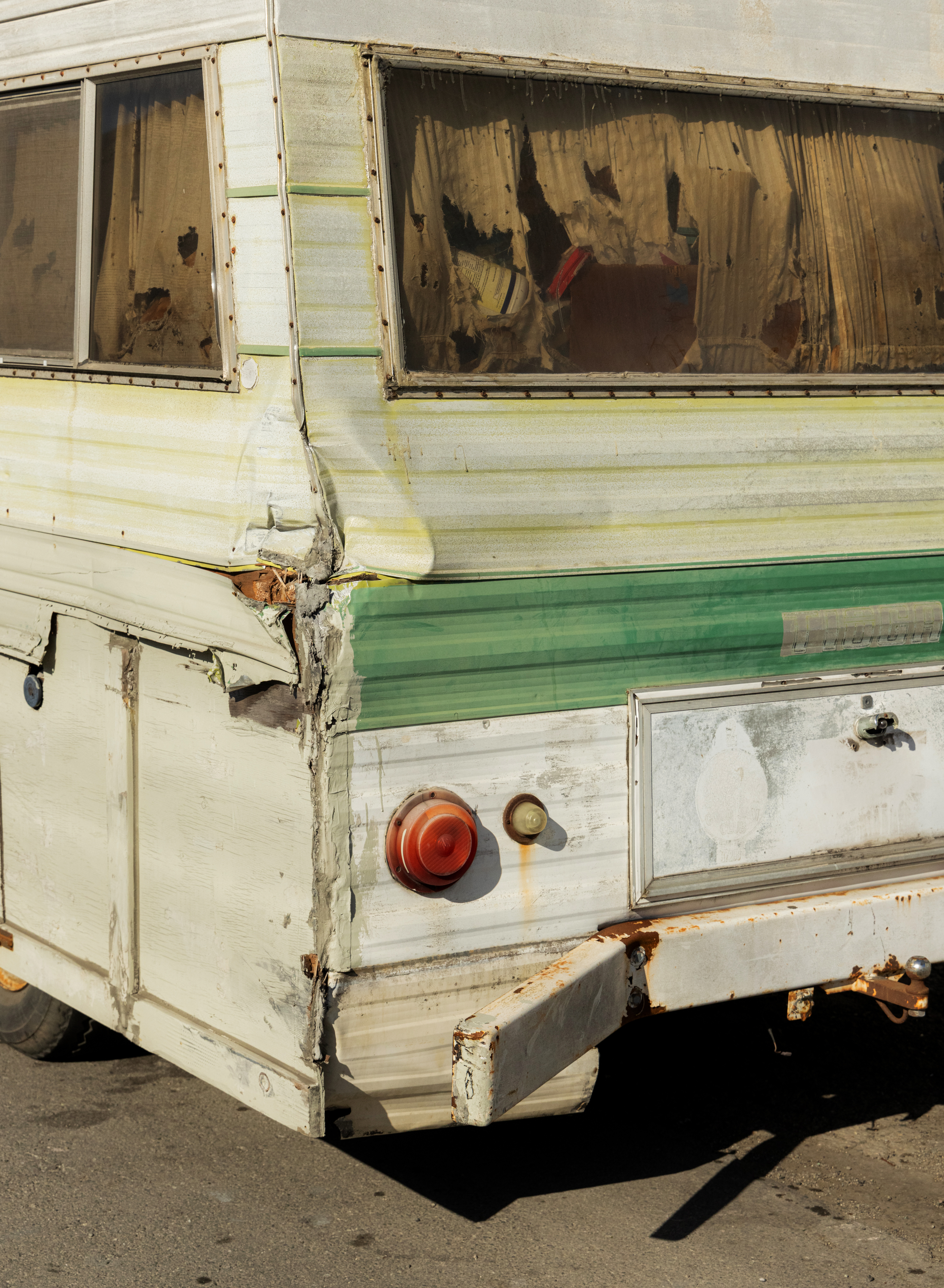 The image shows the damaged rear corner of an old camper, with dented panels, rust, and broken taillights. The windows have torn curtains inside.