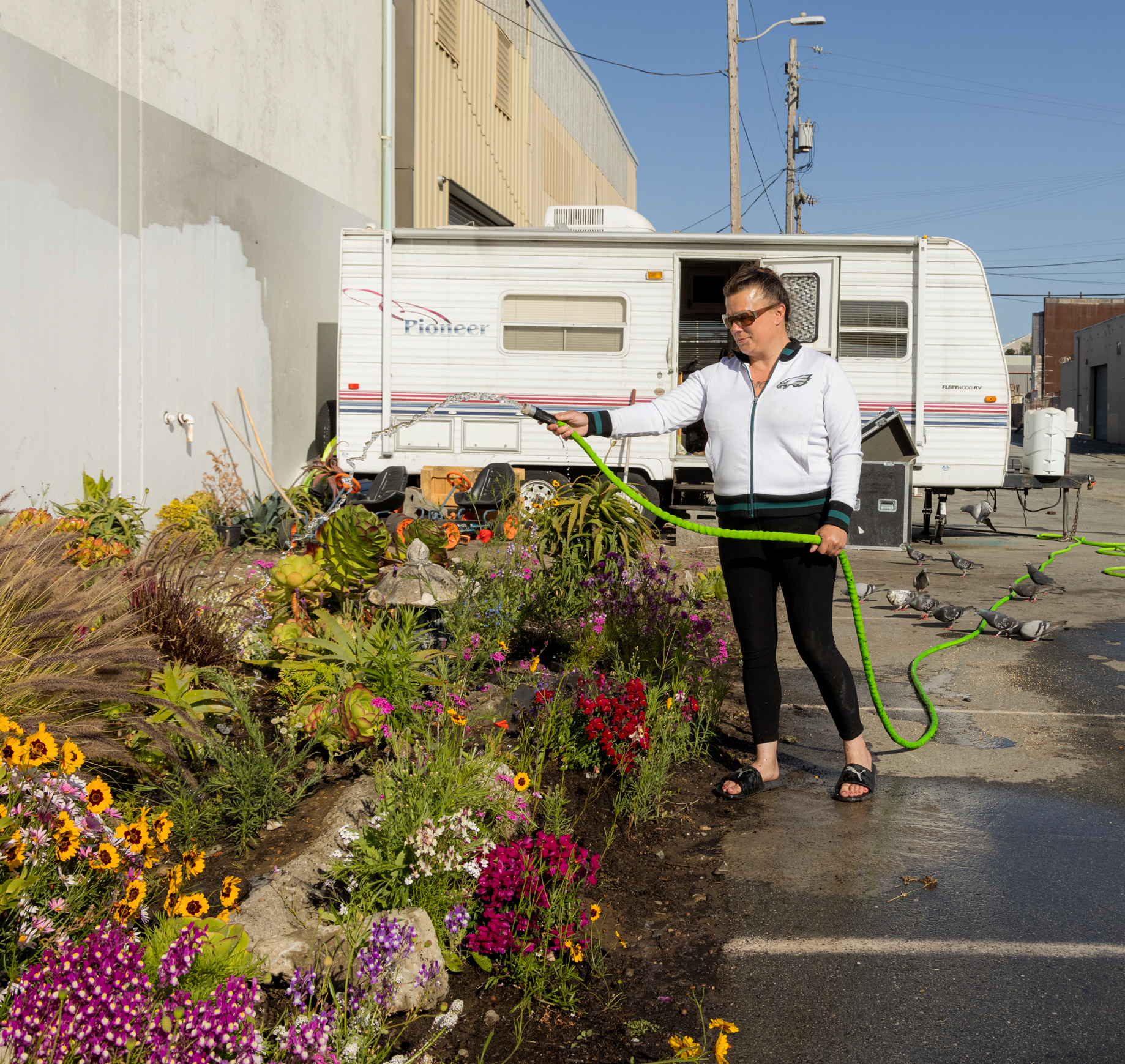 A person waters a colorful garden next to an RV in an industrial area. The garden is filled with various flowers, and the person wears a white jacket and black pants.