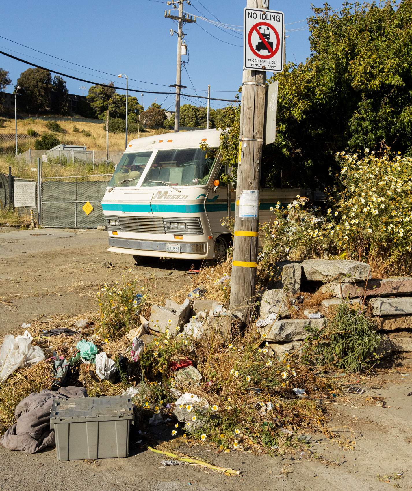 The image shows a weathered RV parked beside a dirt road near a telephone pole with a &quot;No Idling&quot; sign. There is trash and overgrown plants in the foreground.