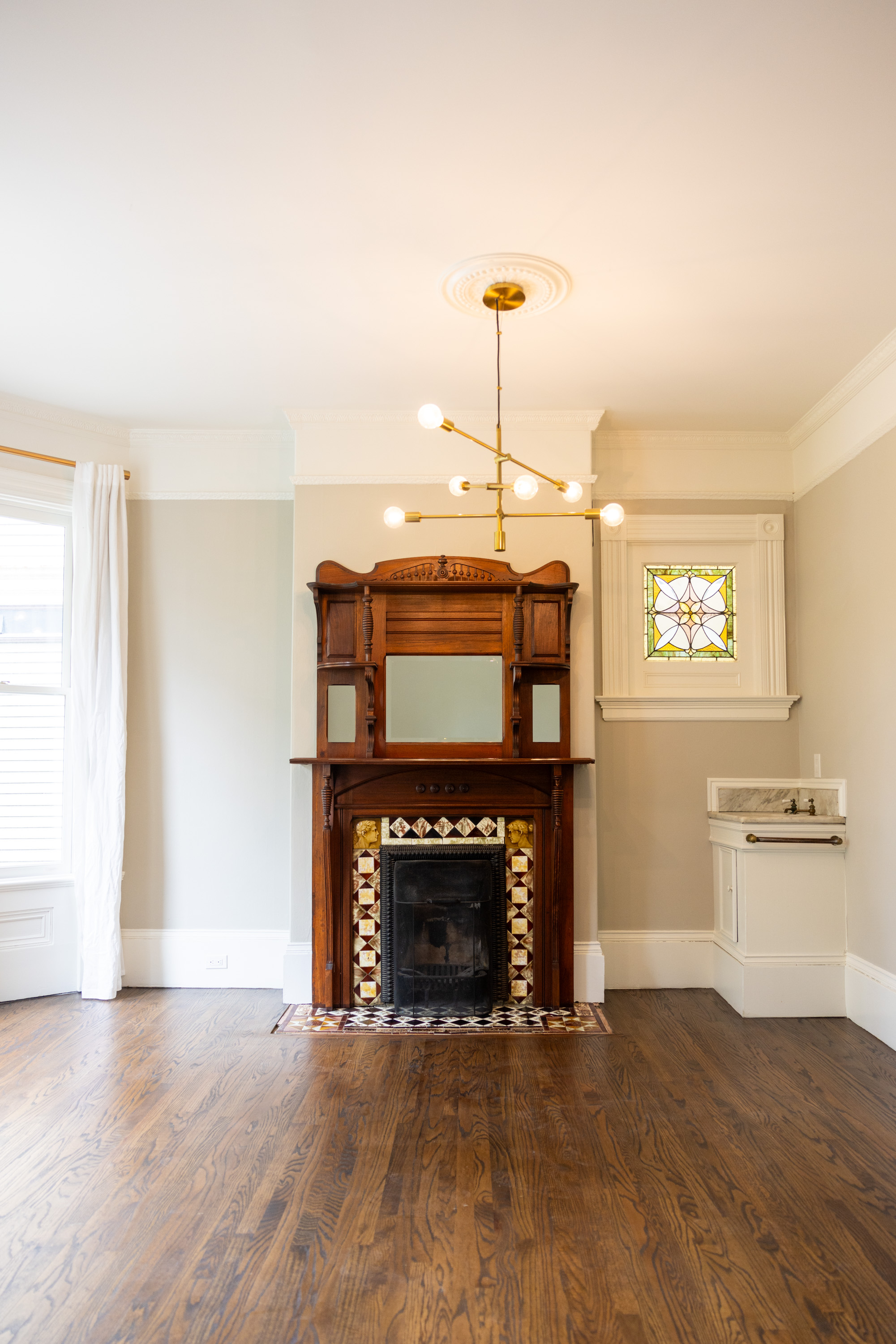 The image shows a room with wooden floors, a decorative wooden fireplace with an ornate mirror, a modern chandelier, stained glass window, and a small white sink unit.
