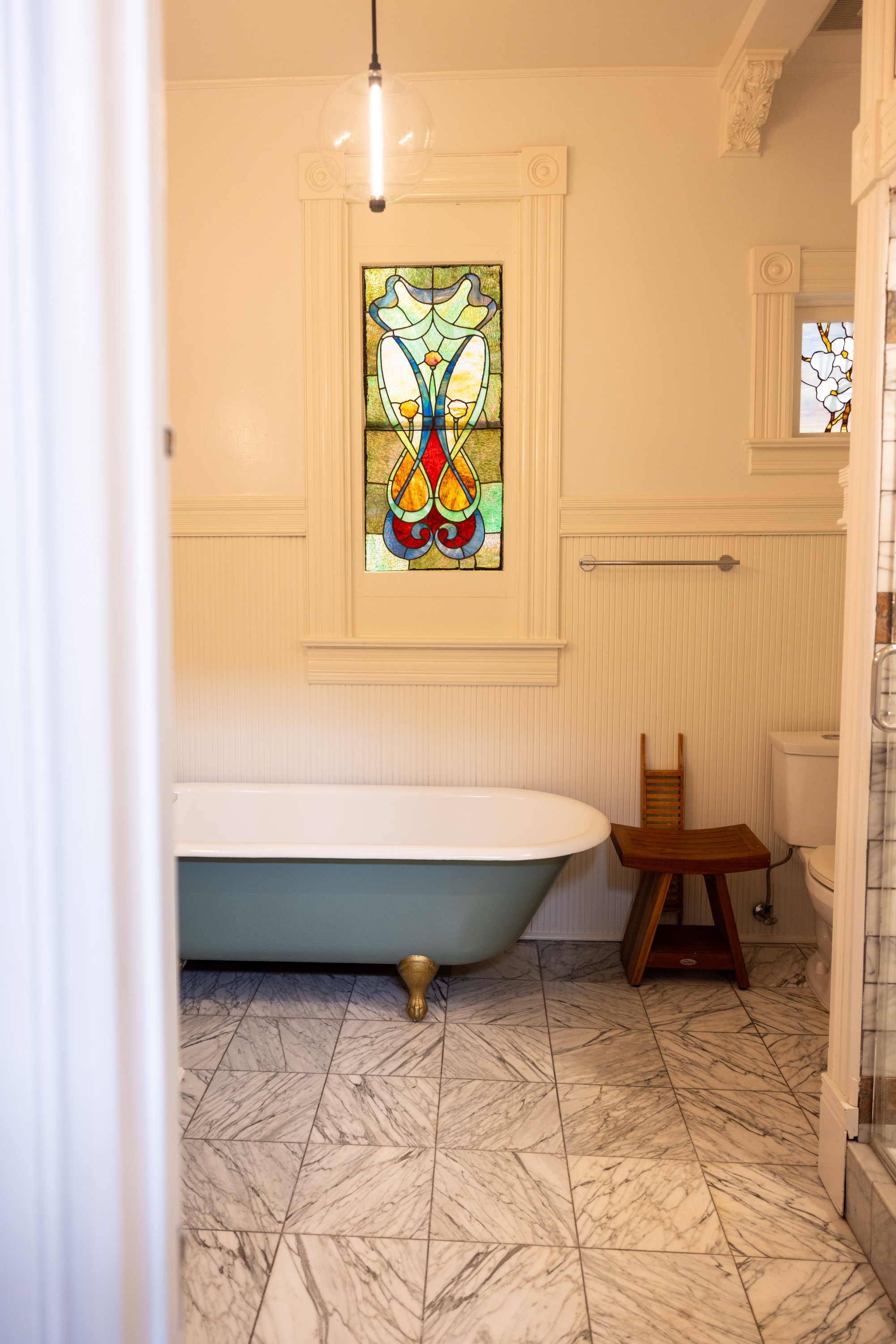 The image features a bathroom with a vintage bathtub on golden legs, marble floor, stained glass window, and a wooden chair beside a white toilet under soft lighting.