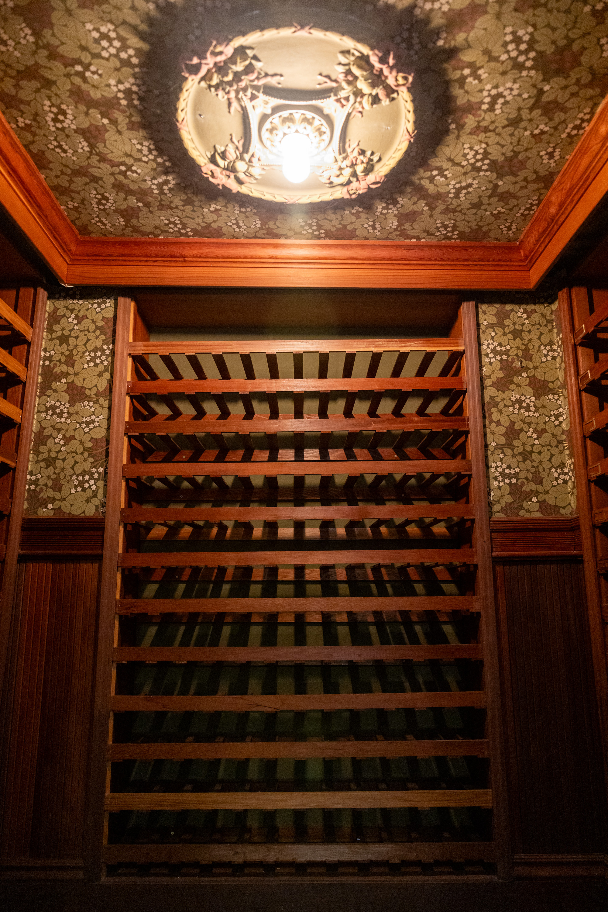 The image shows ornate ceiling lights and patterned wallpaper above empty wooden wine racks in a dimly lit room.