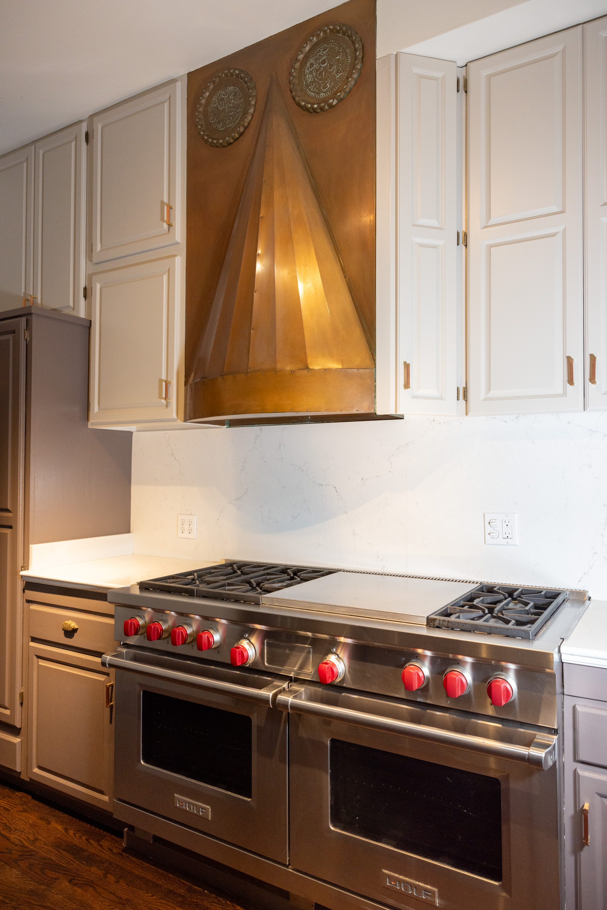 The image shows a modern kitchen with a stainless steel stove featuring six red knobs and dual ovens. Above, there's a unique copper range hood surrounded by white cabinets.