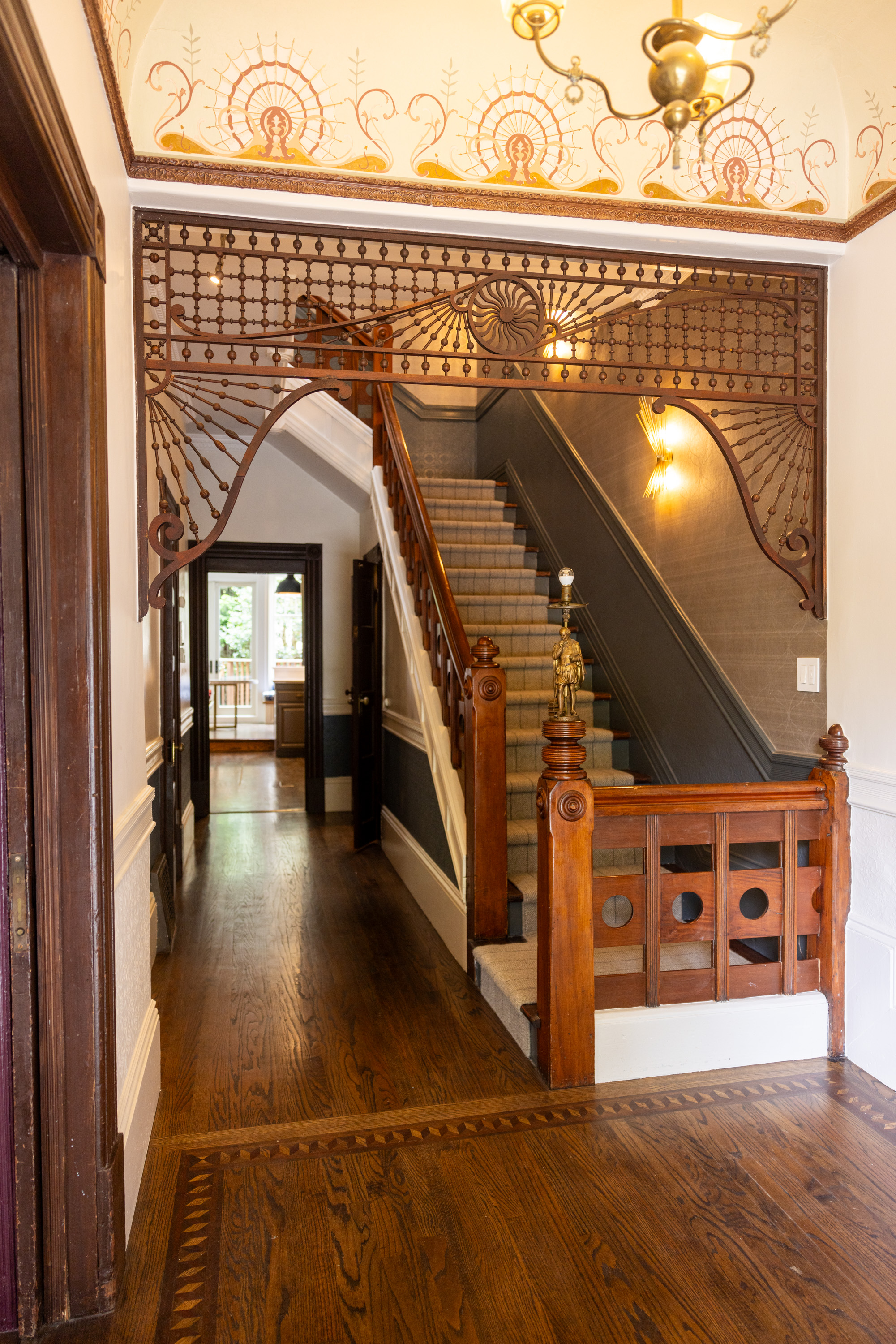 The image shows an ornate, narrow hallway featuring a wooden staircase with detailed railings and newel posts, with patterned wallpaper and decorative stenciling on the walls.