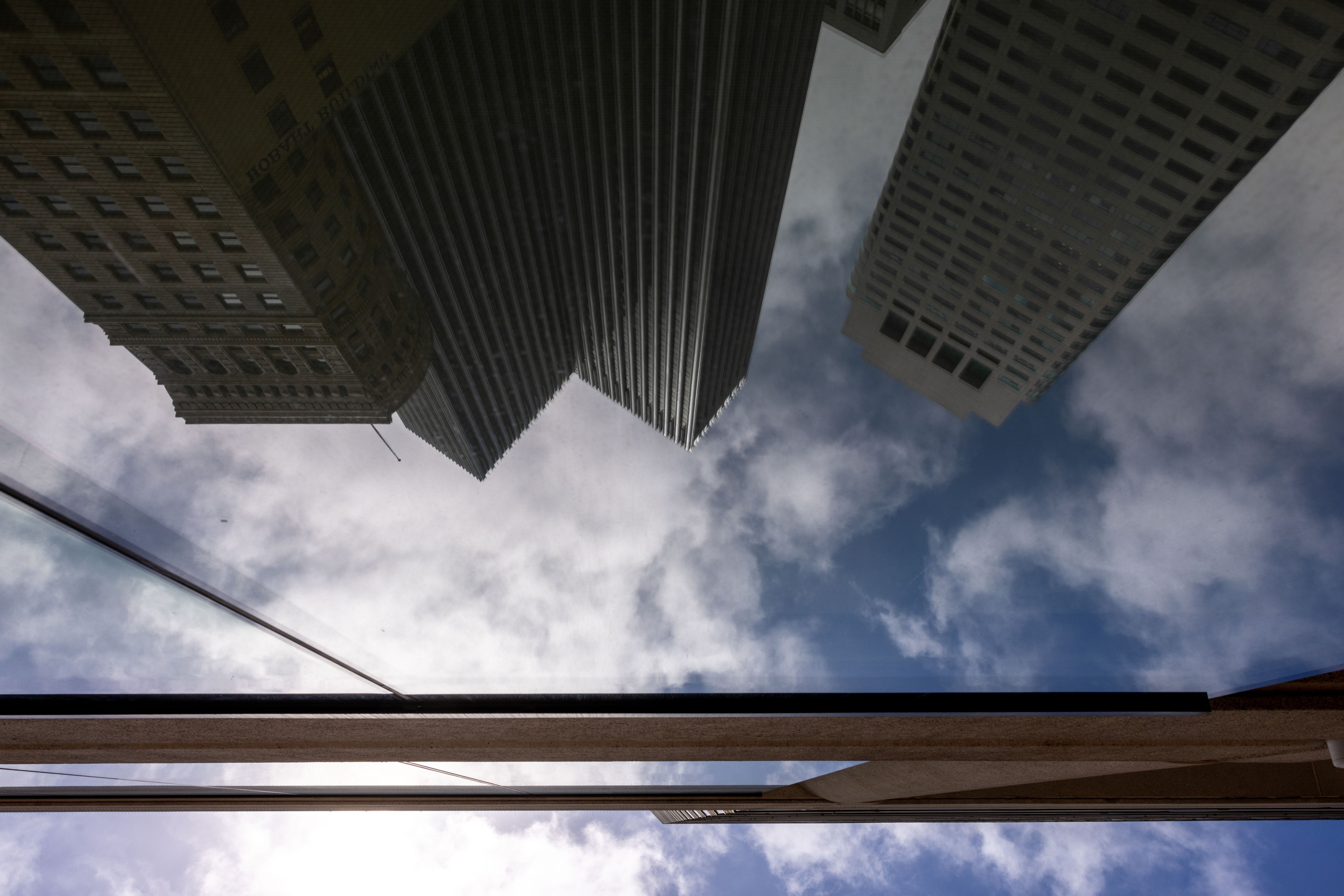 The image shows a reflection of tall buildings in a glass surface, with a partly cloudy sky and blue patches visible above.