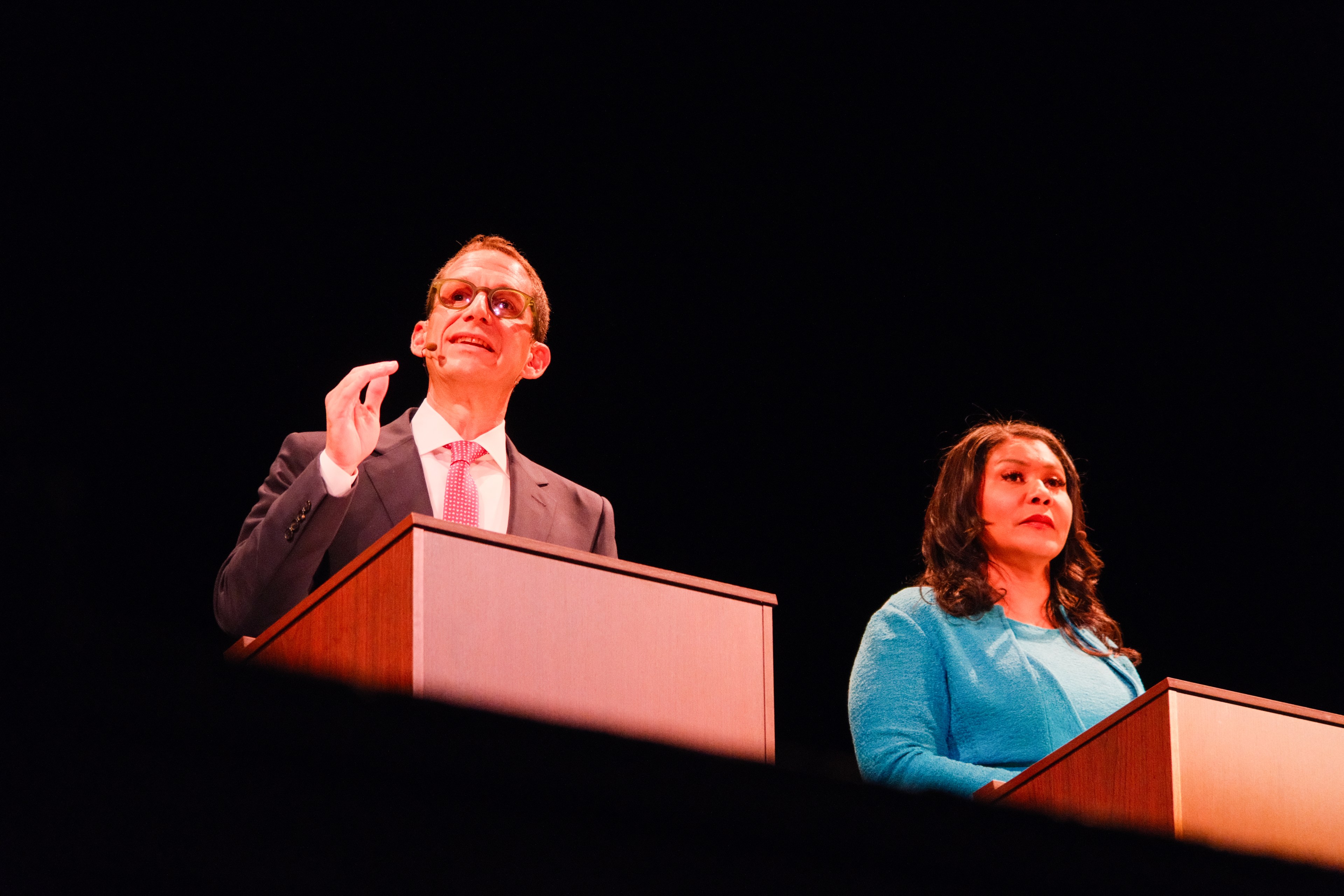 Two people, a man in a suit and tie, and a woman in a blue outfit, are standing behind podiums on a stage, speaking, against a black background.