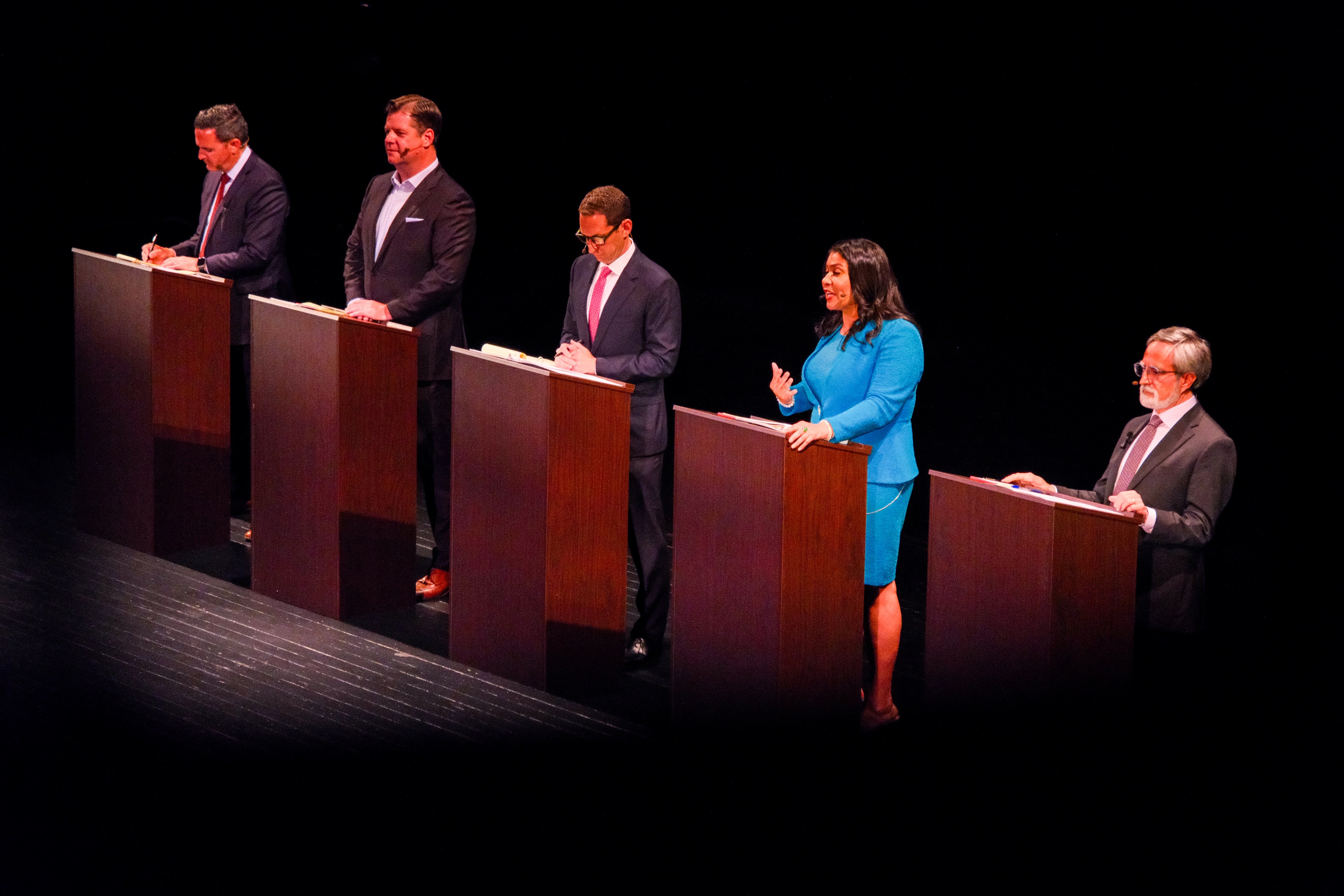 Five people stand behind podiums on a stage, participating in what appears to be a debate or panel discussion.