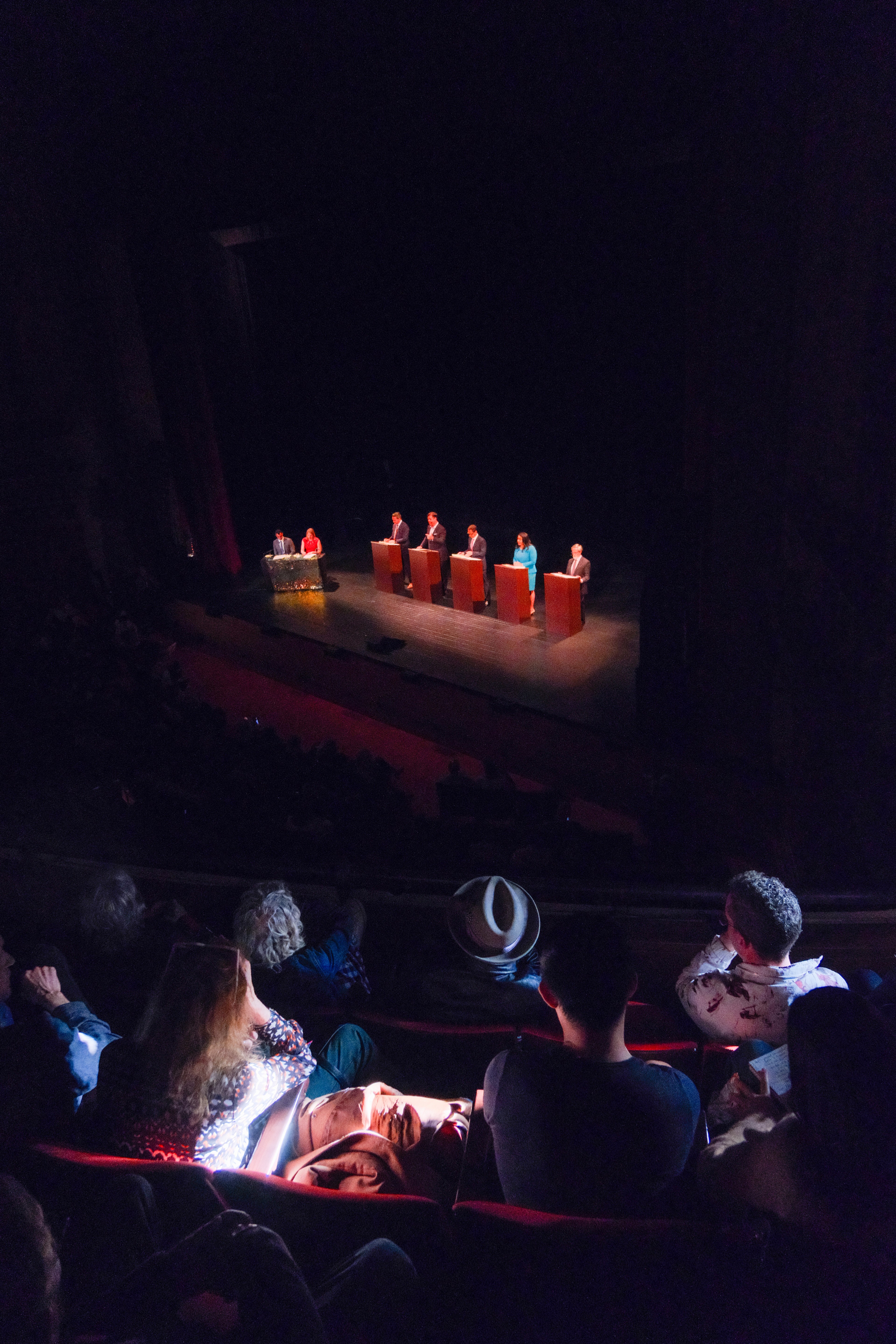 An audience in a theater watches six people at podiums on a stage. The setting is dimly lit, with the focus on the stage and the audience seated in semi-darkness.