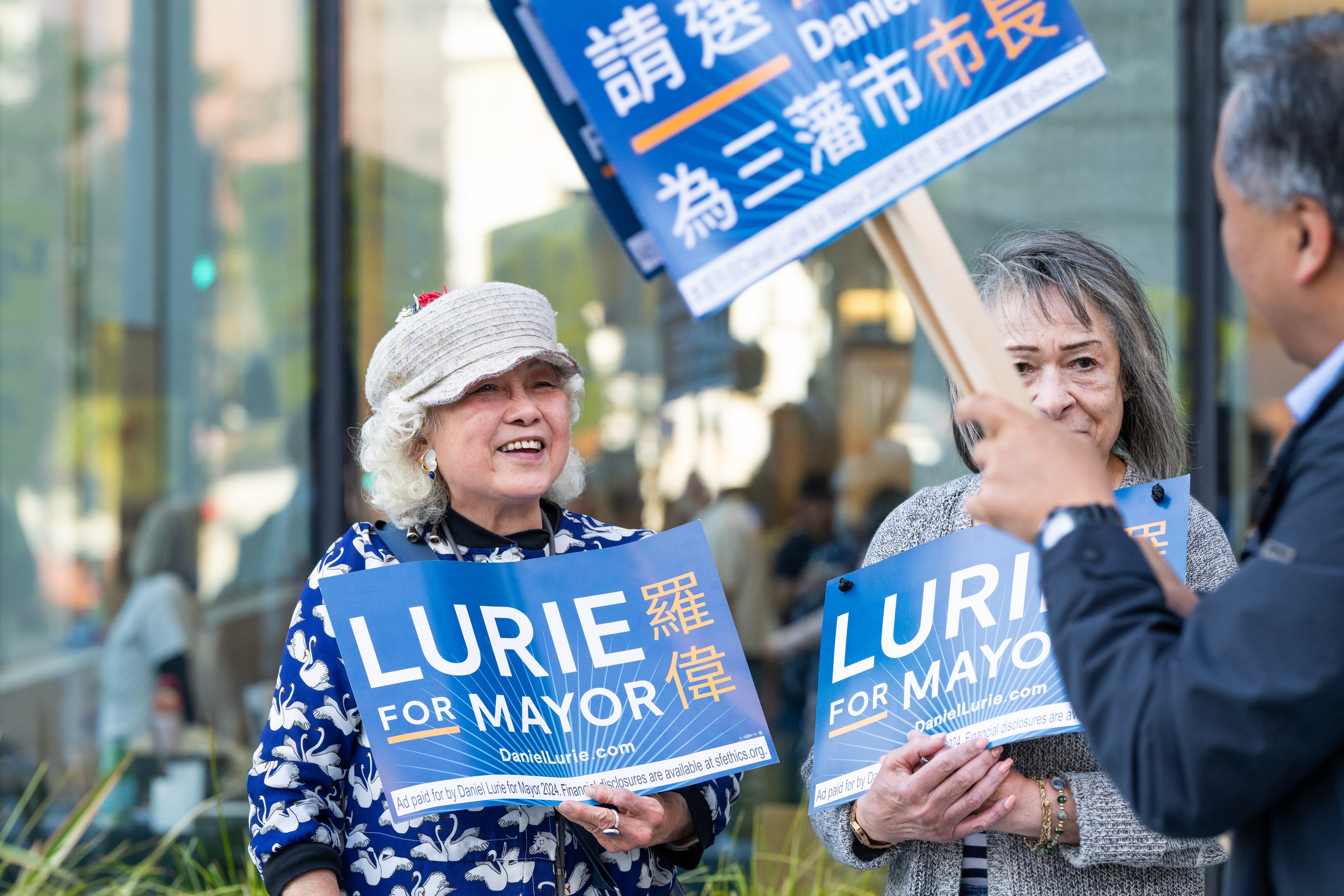 People seen from the waist up, outside in a city street, hold blue campaign signs reading &quot;LURIE.&quot;