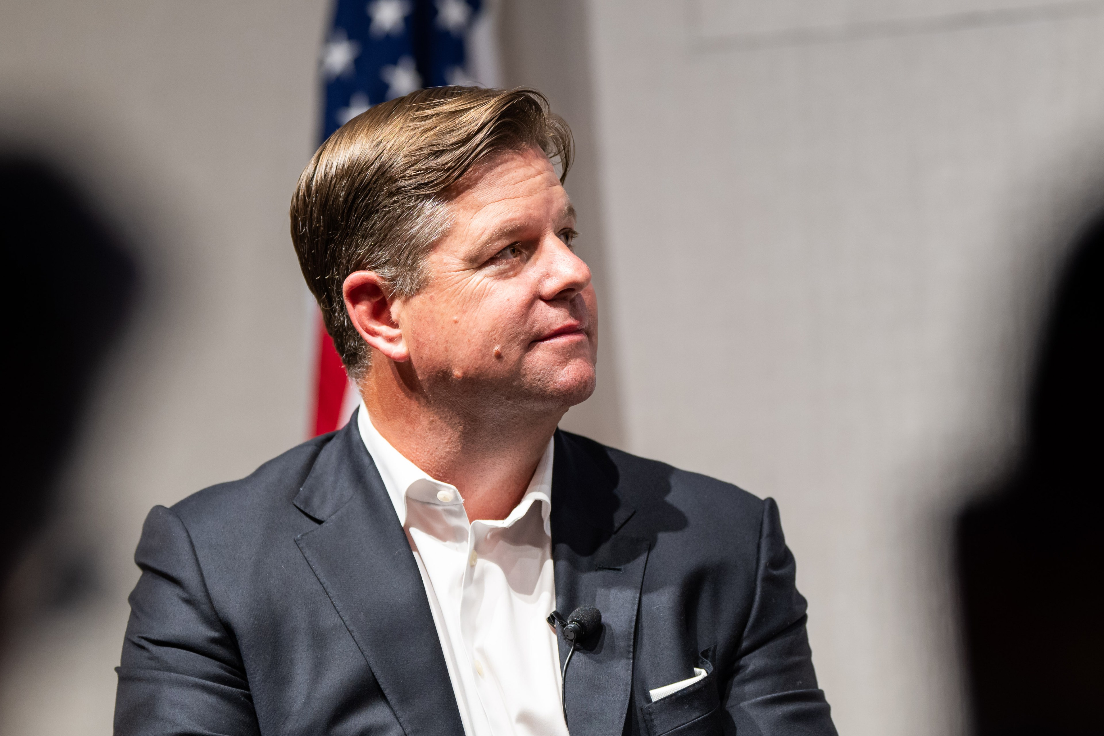 A man in a suit looks to the side, sitting with an American flag partially visible in the background. He has a microphone clipped to his shirt.